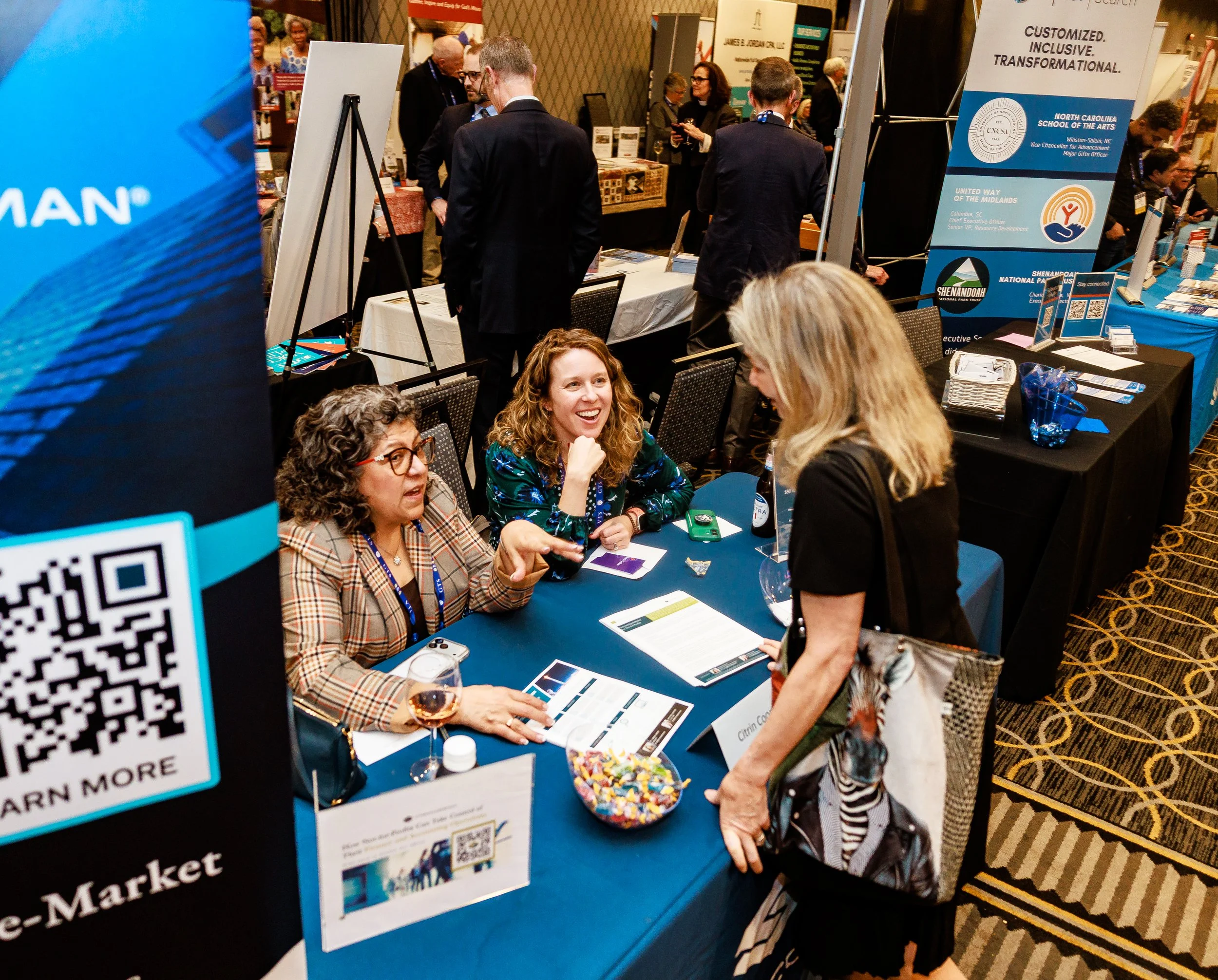 Three women at a conference booth engaging in conversation, with some smiling, on a blue table with informational materials, a bowl of candy, and a glass of wine, surrounded by banners and other booths in the background.