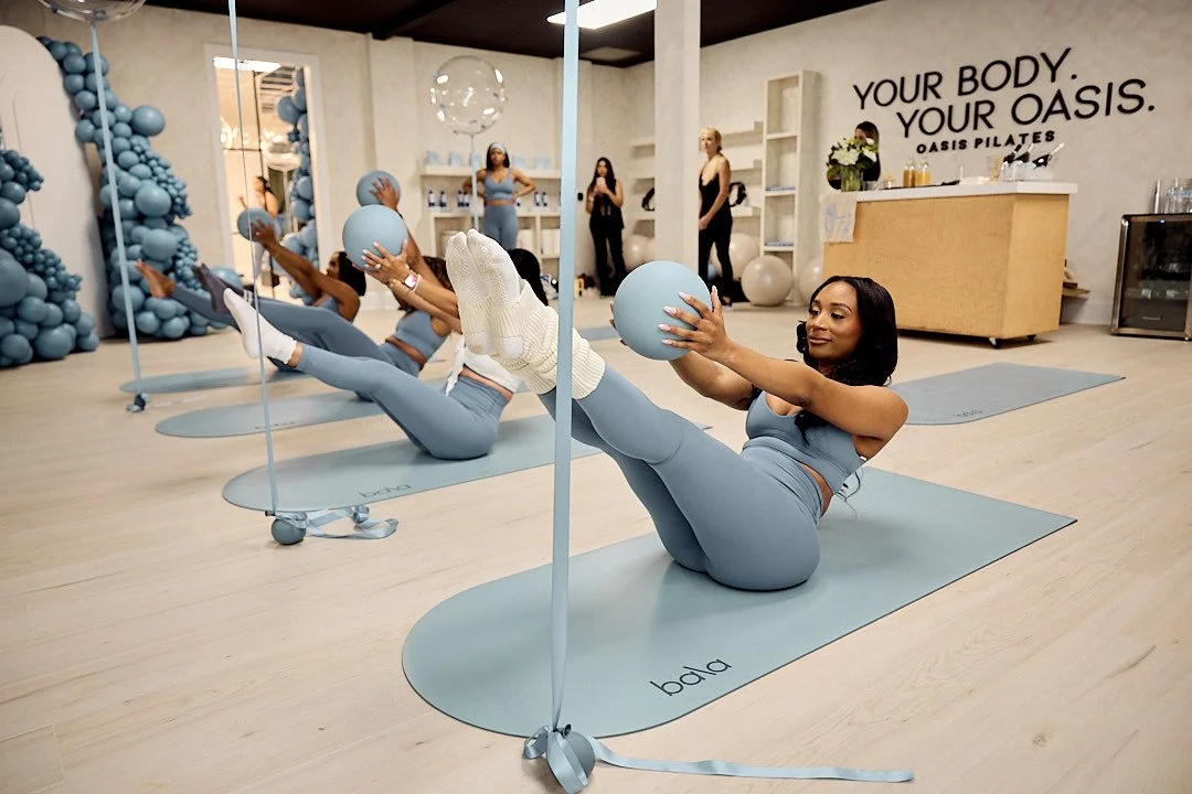 Women participating in a Pilates class in a studio, doing leg raises with exercise balls, with a sign on the wall reading 'Your Body, Your Oasis'.