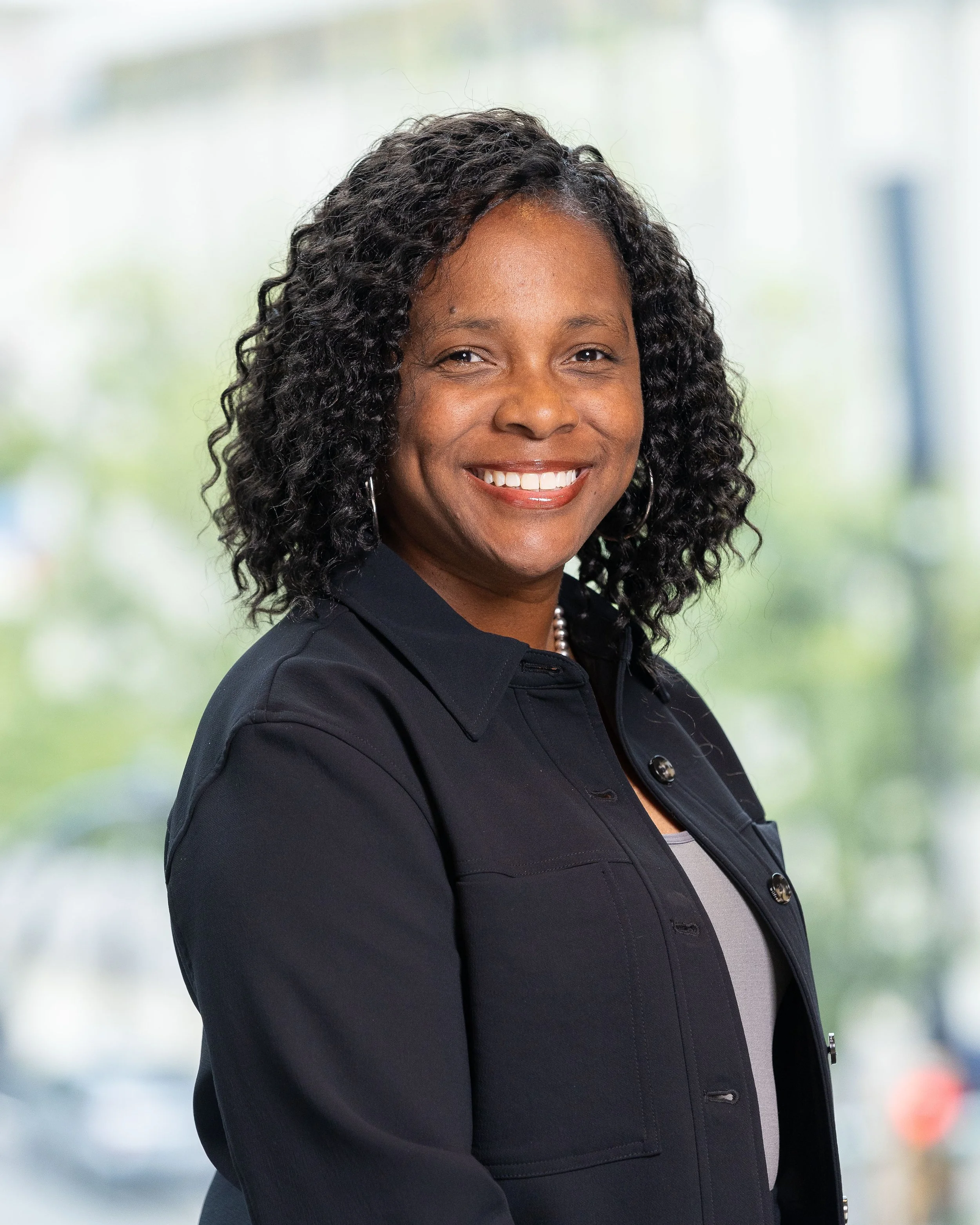 Portrait of a smiling woman with curly hair, wearing a dark blazer, standing against a blurred outdoor background.