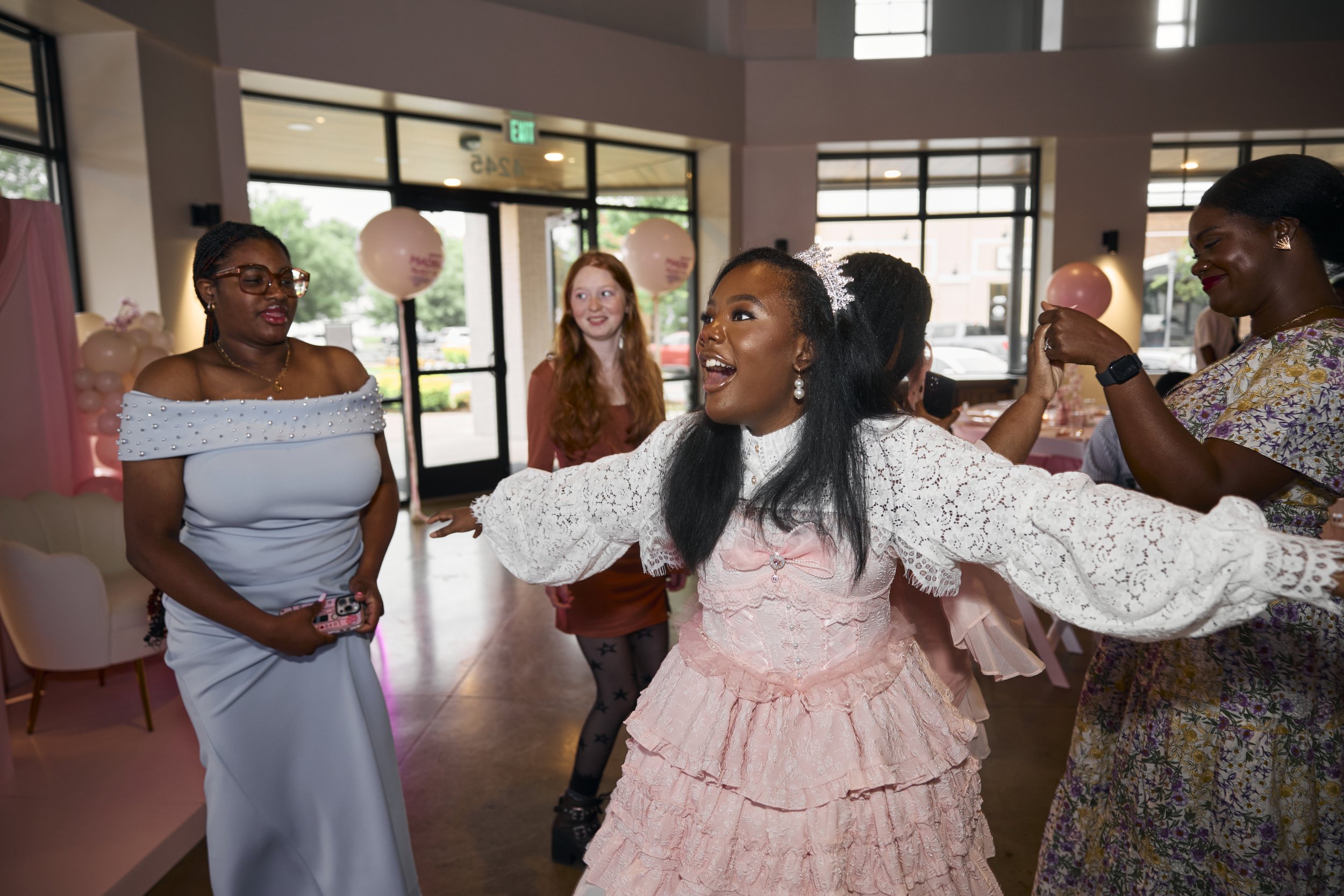 Group of women at a celebration, with one woman in a pink dress dancing joyfully while others watch and smile inside a decorated venue.