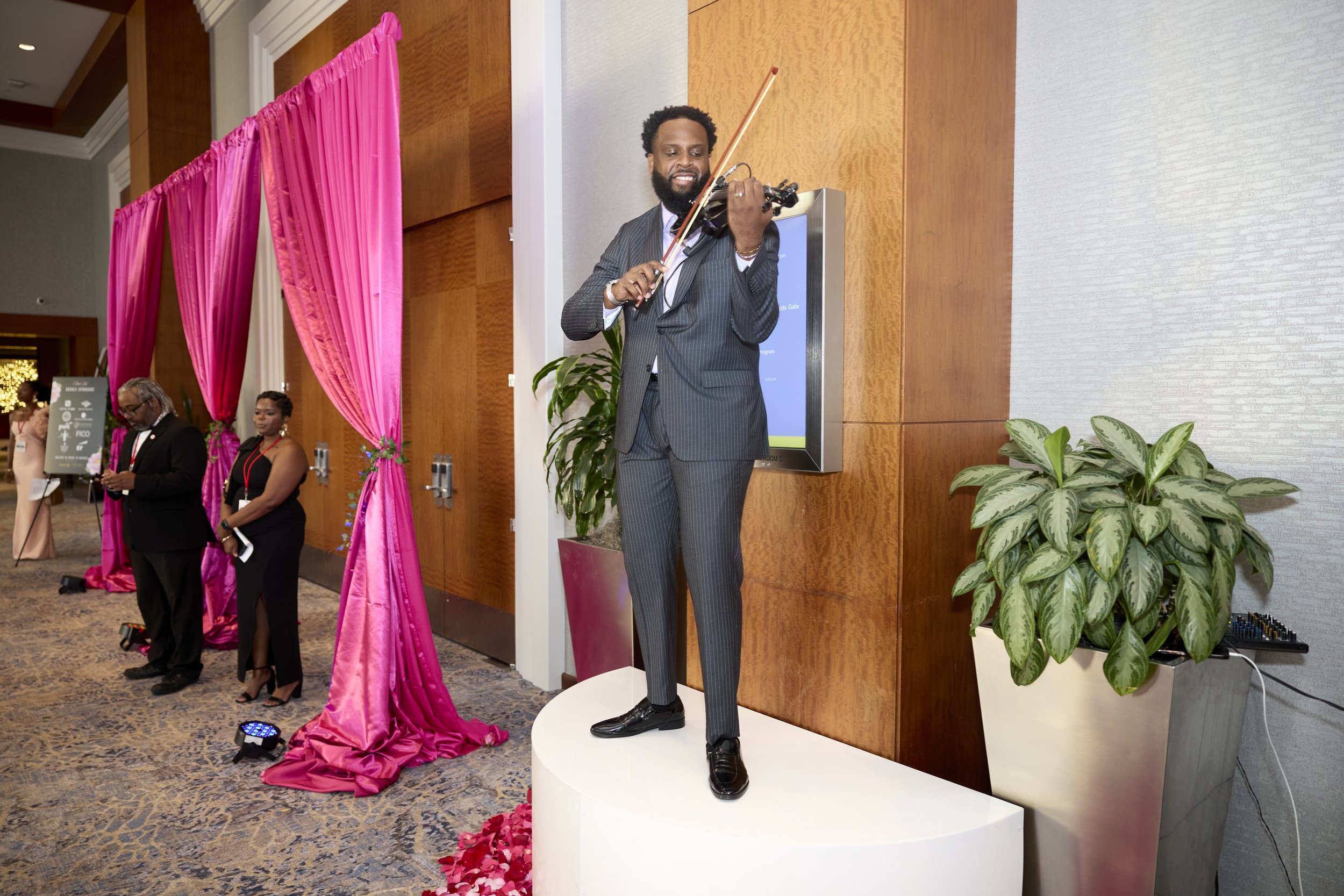 Man in a dark pinstripe suit standing on a white platform, playing a violin at an indoor event with pink draping and potted plants.