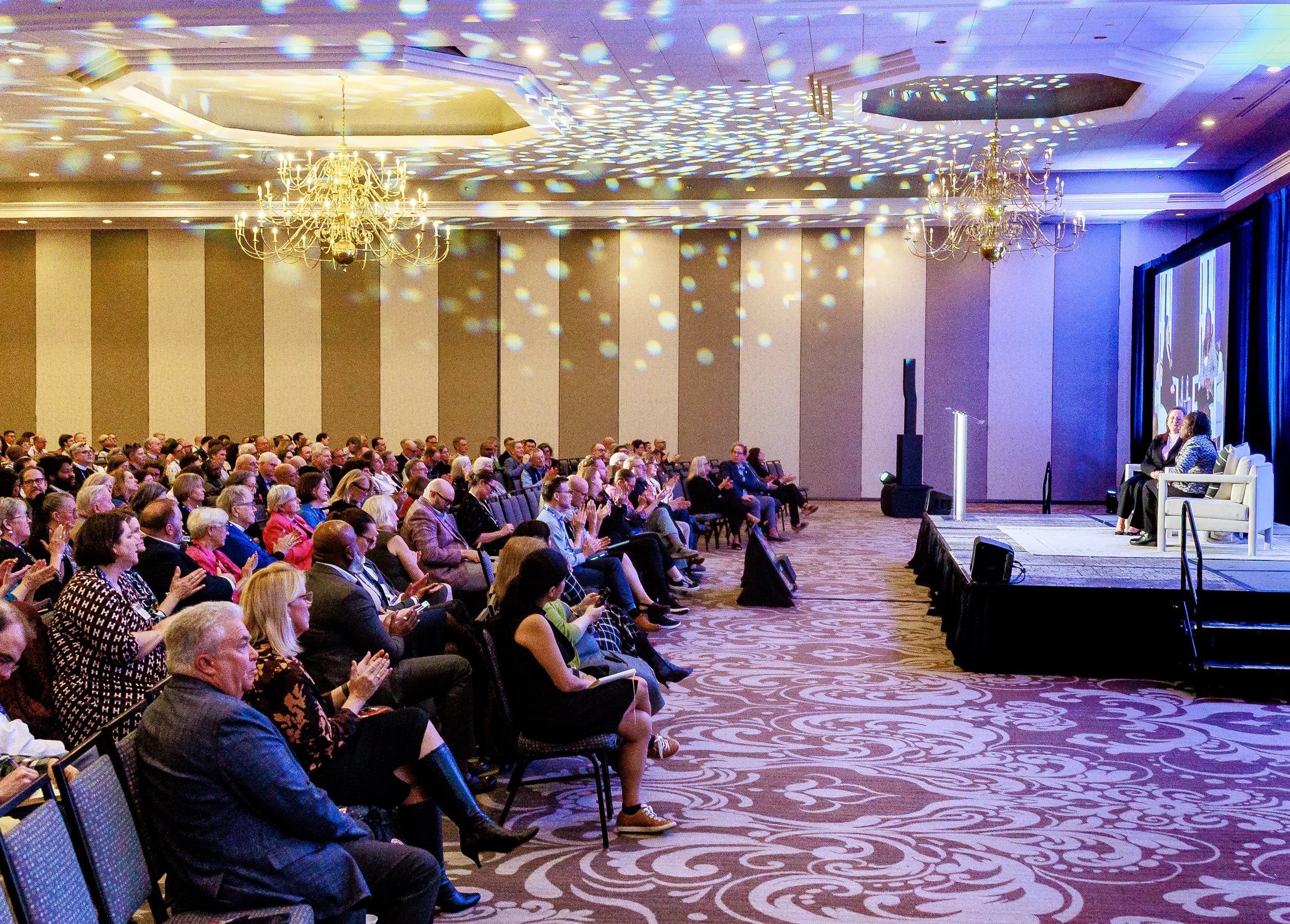 Audience attending a panel discussion in a large, elegant conference room with chandeliers and patterned carpet, facing a stage with three speakers seated in white chairs and a large screen behind them.