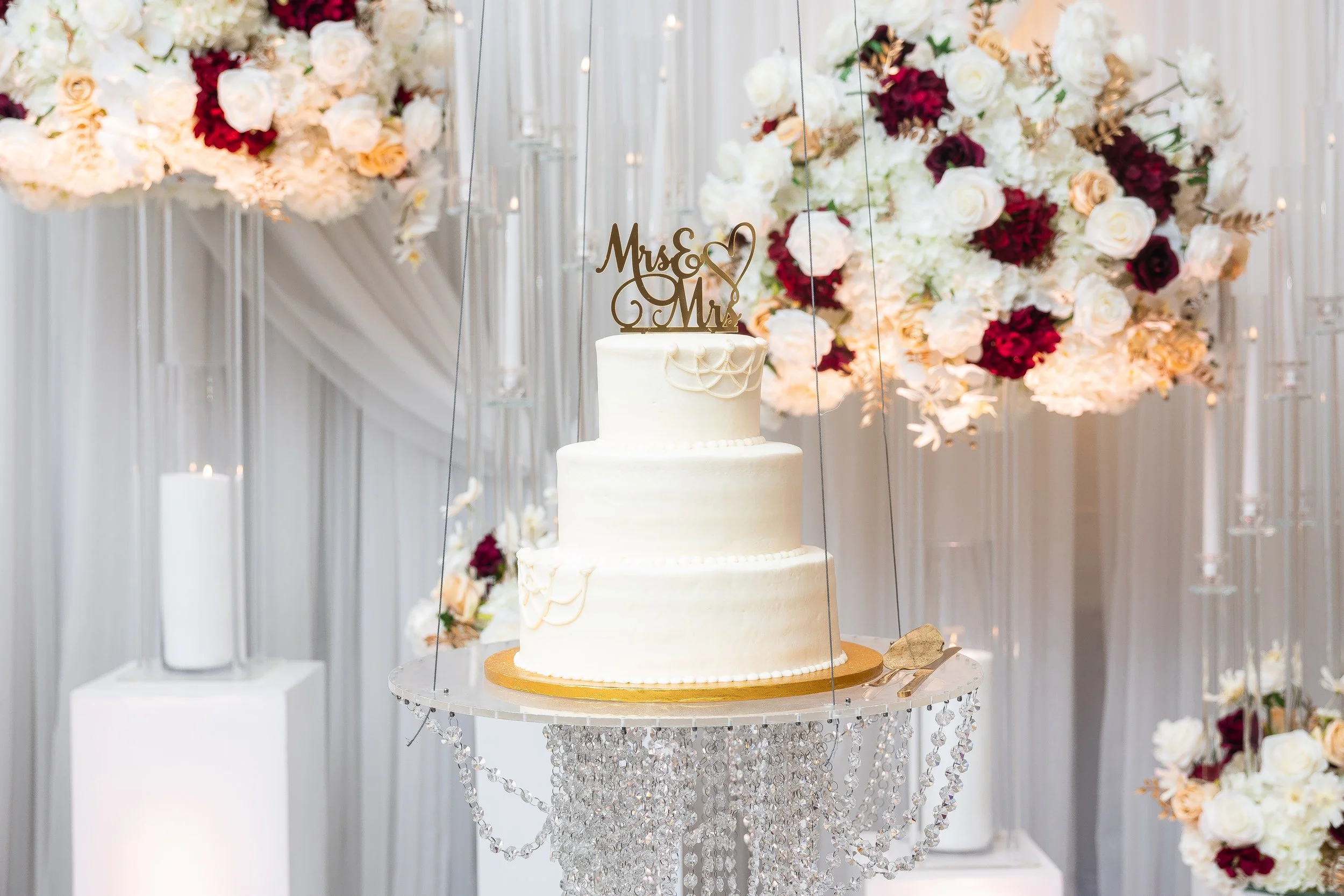 A three-tier white wedding cake with gold 'Mrs & Mr' topper, decorated with white and gold accents, sits on a crystal-encrusted stand in front of a backdrop of white and red flowers.