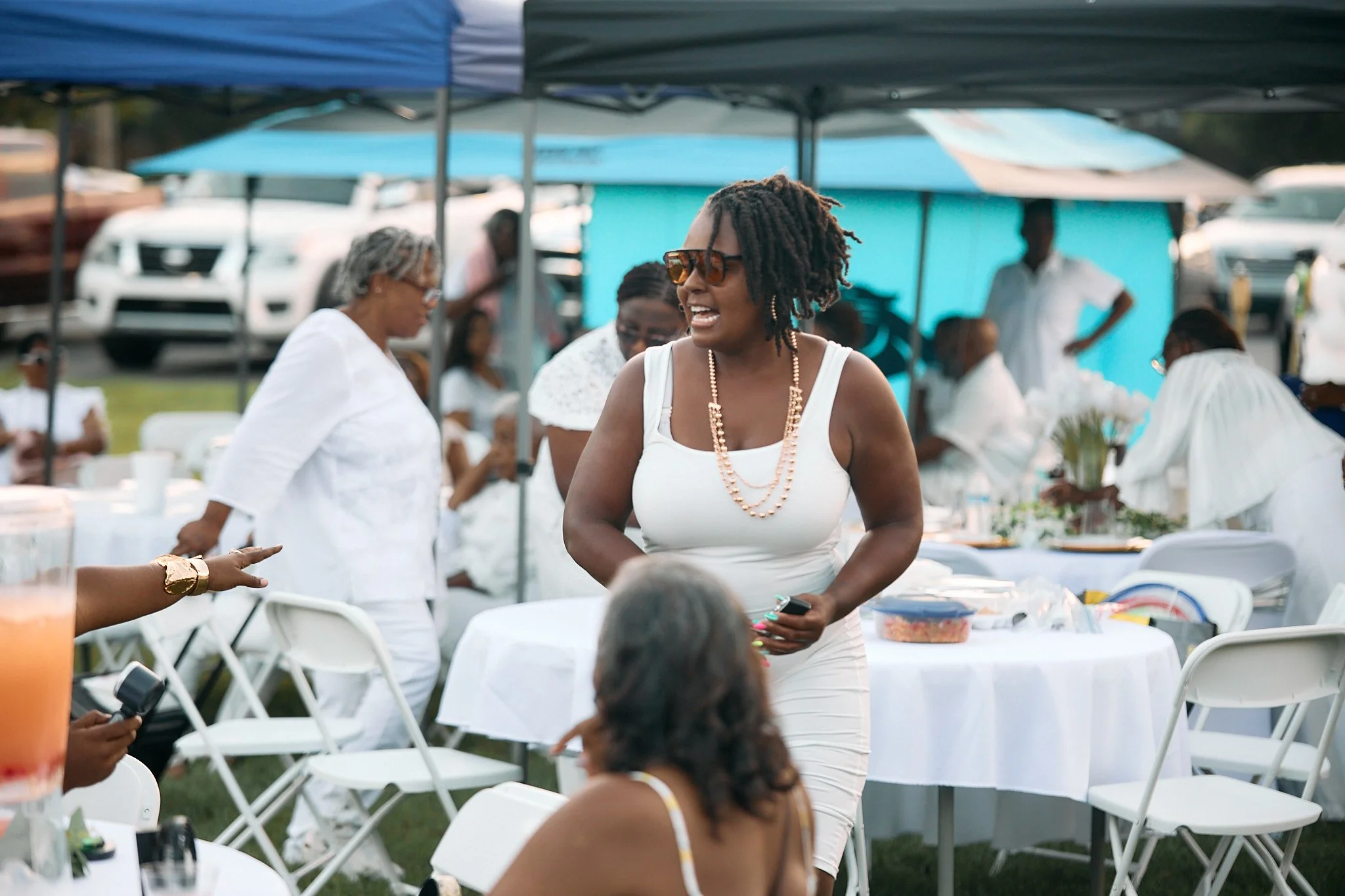 A woman in a white dress with curly hair, sunglasses, and a pearl necklace is standing and talking at an outdoor event under a tent with white chairs and tables around. Other people are gathered in the background.