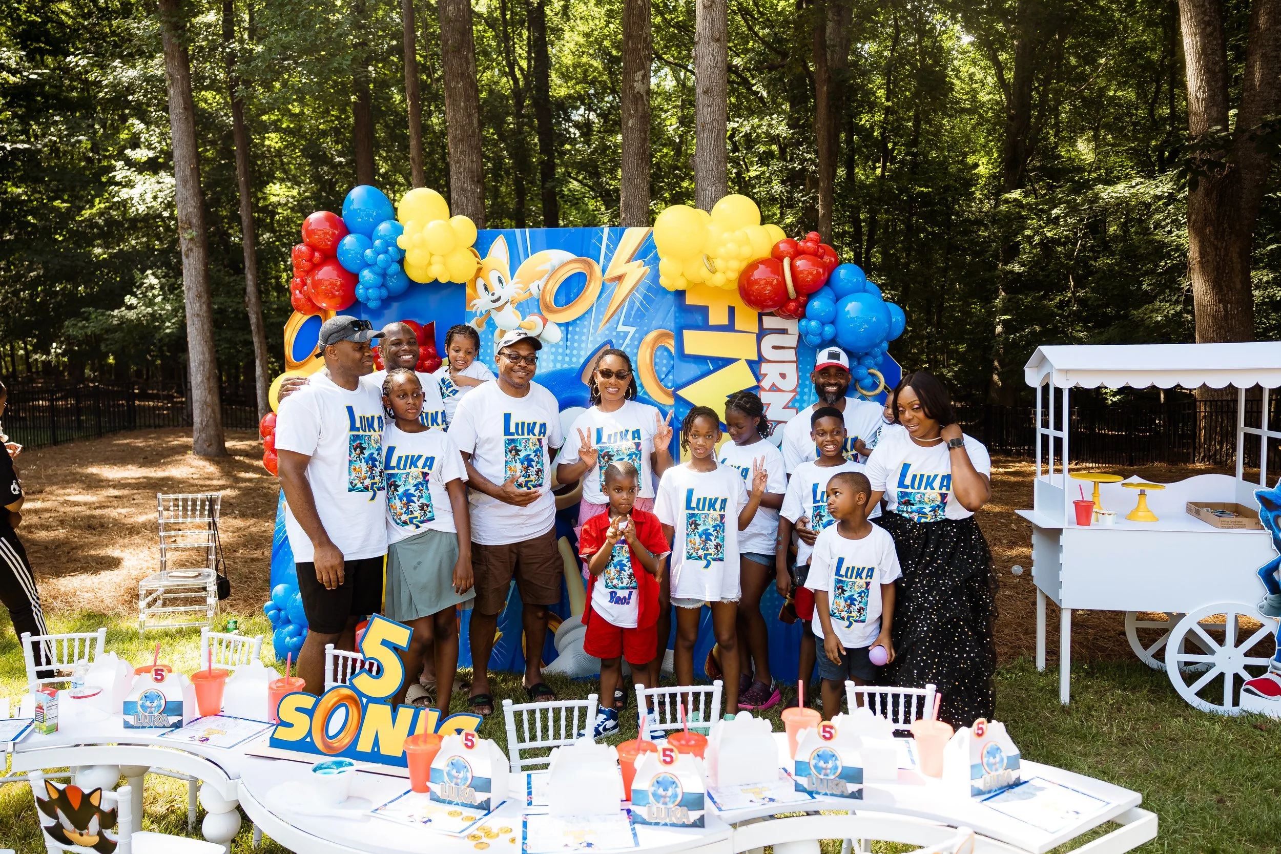 Children and adults at a Sonic the Hedgehog themed birthday party outdoors, with a decorated backdrop, balloons, and a table with party favors.