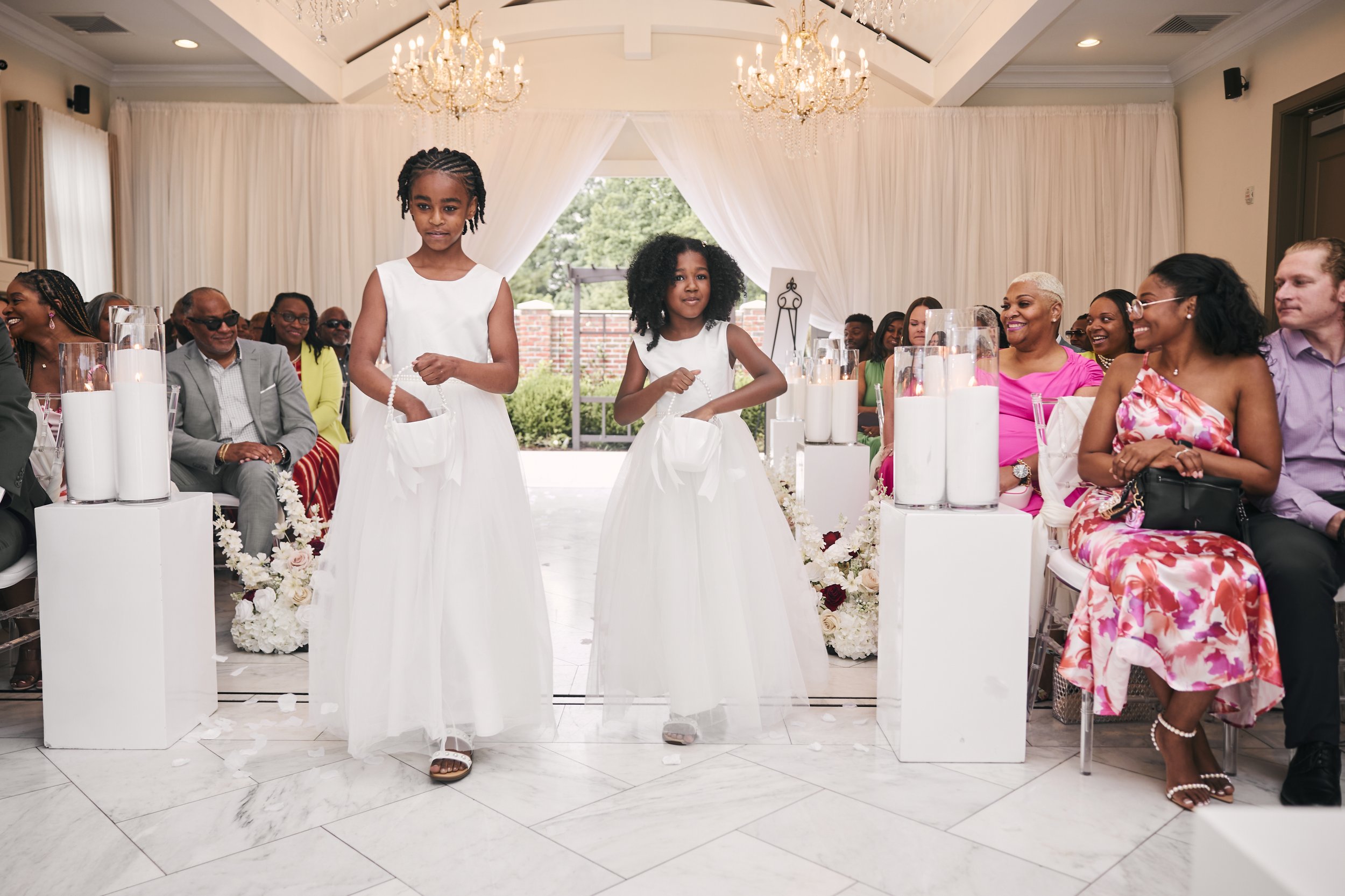 Two young girls in white dresses are walking down the aisle of a decorated venue during a wedding ceremony, with seated guests smiling and watching.