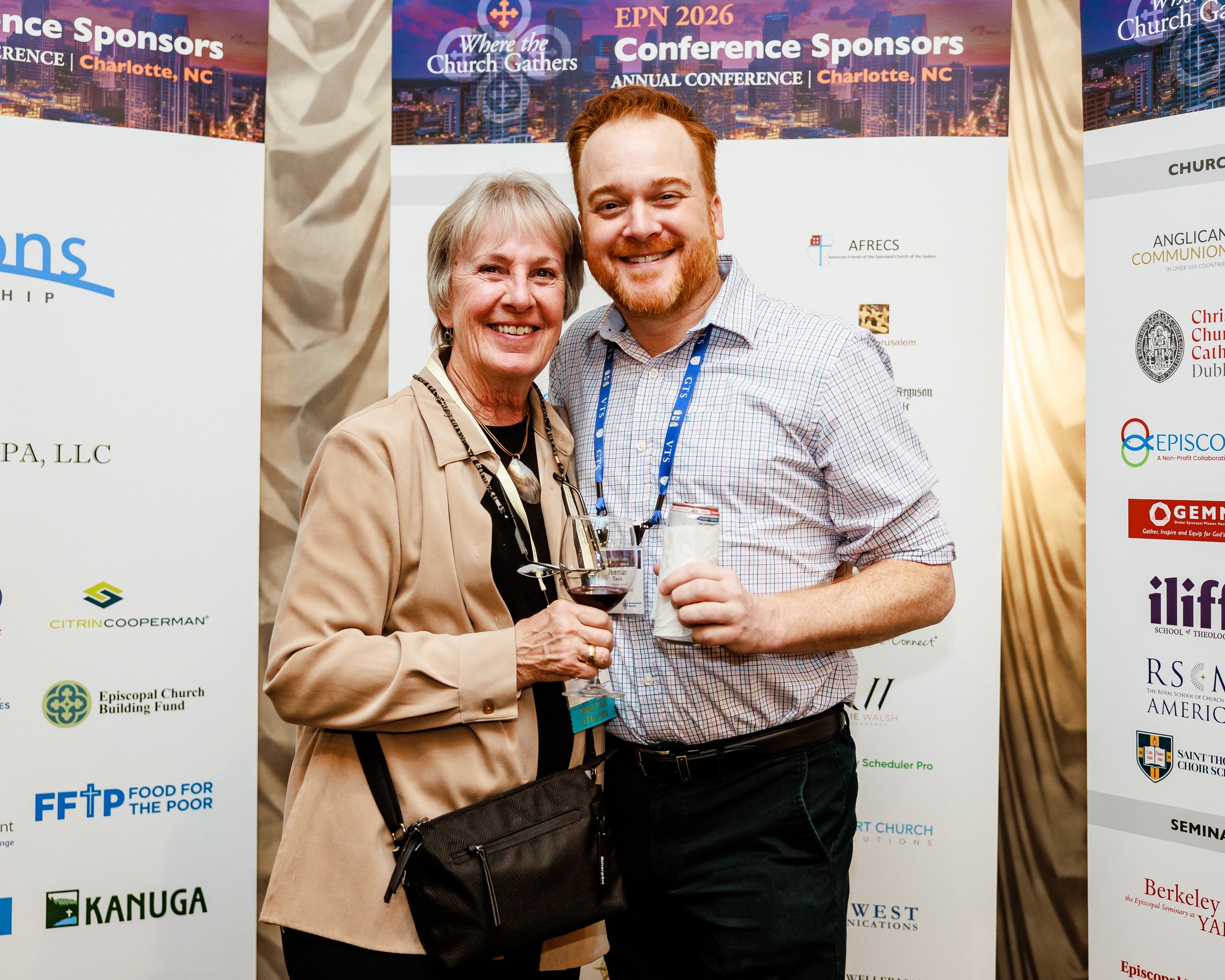 A woman and a man smiling and holding drinks at a conference. The woman has gray hair and is wearing a beige blazer and black shirt. The man has red hair and a beard, wearing a checkered shirt and black pants. They are standing in front of a conferen