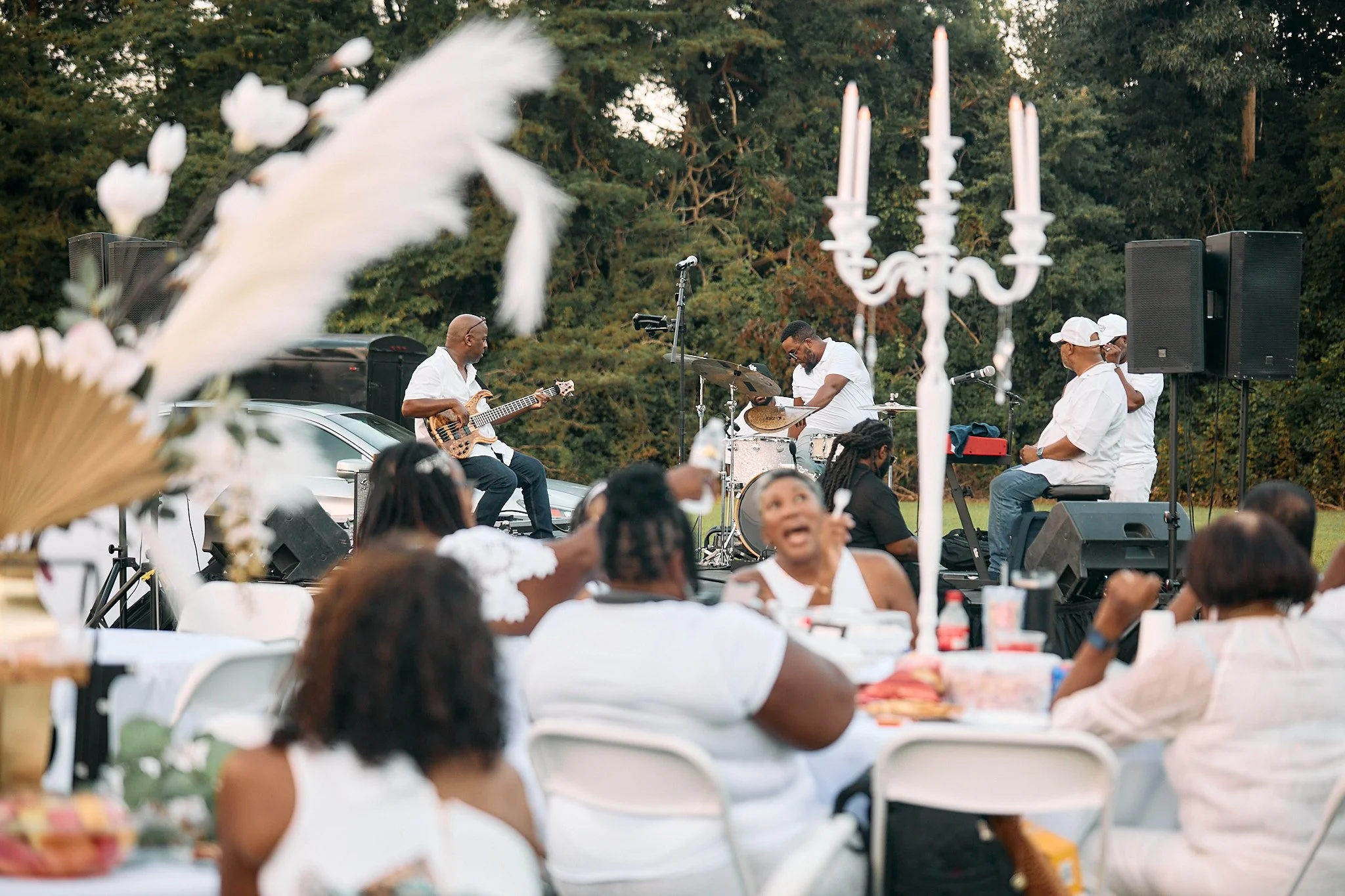 Outdoor musical performance with a band of four musicians playing instruments and several audience members seated at tables, some of whom are smiling and enjoying the event, with trees in the background.