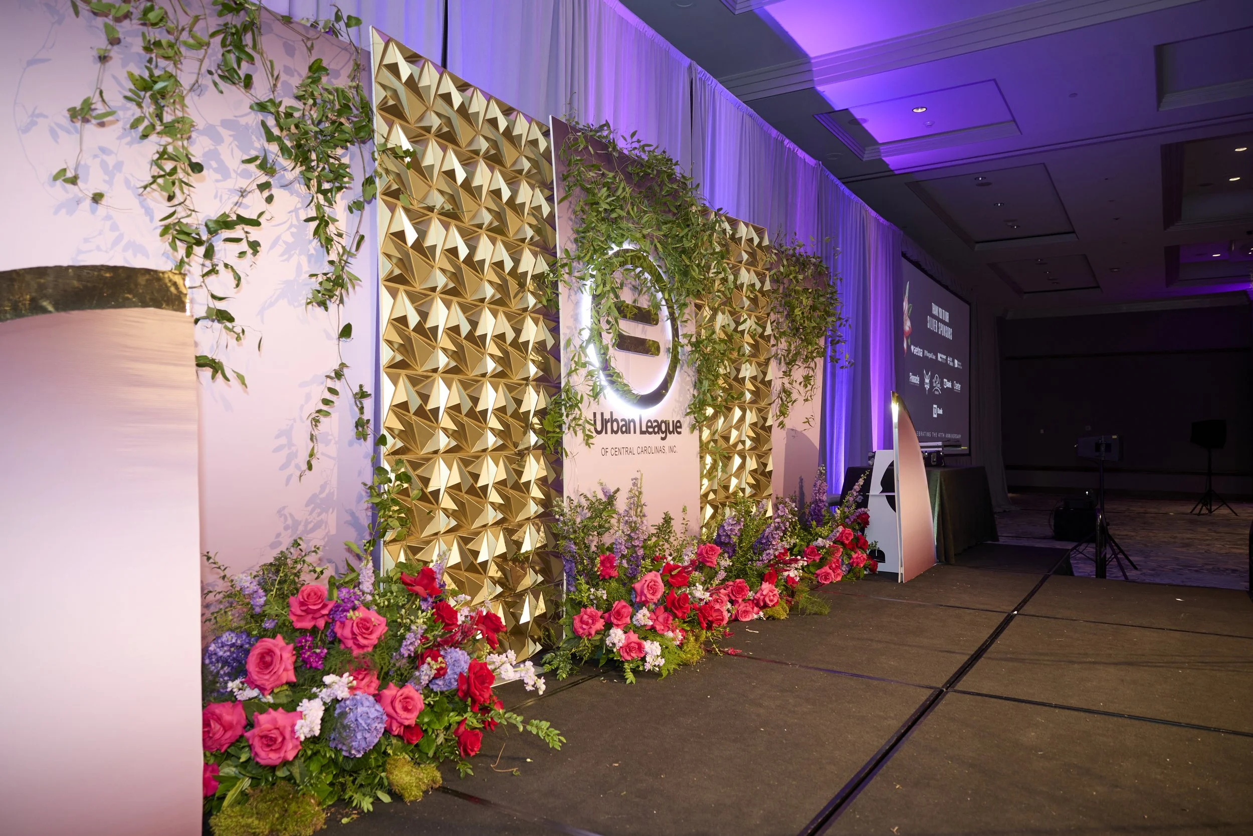 Event stage decorated with gold geometric panels, floral arrangements with pink and purple flowers, and a sign for Urban League of Central Carolinas, Inc., with purple uplighting and curtains.