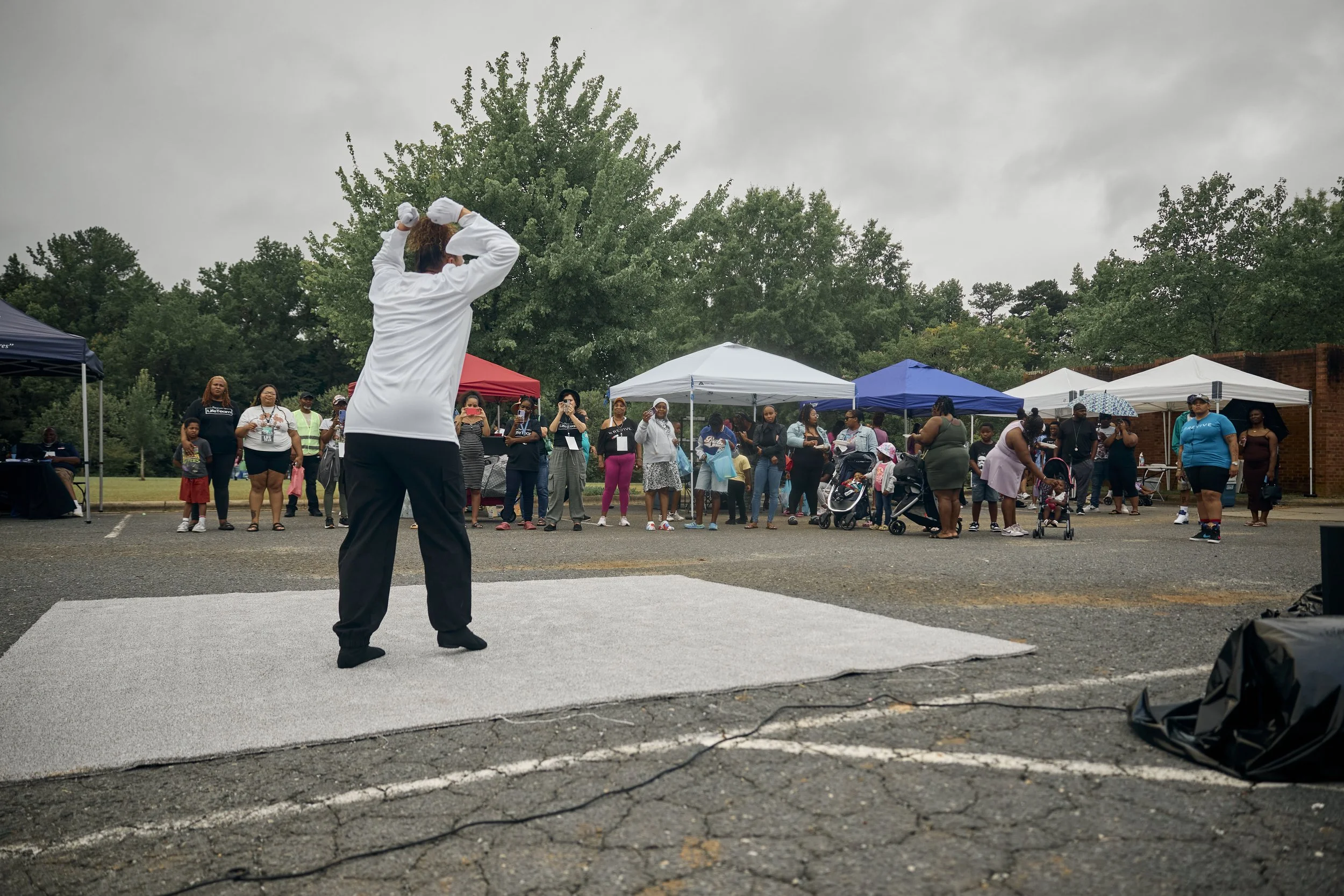 A person stands on a small stage or platform, holding a microphone and facing a group of people gathered outdoors under tents, with trees and overcast sky in the background.