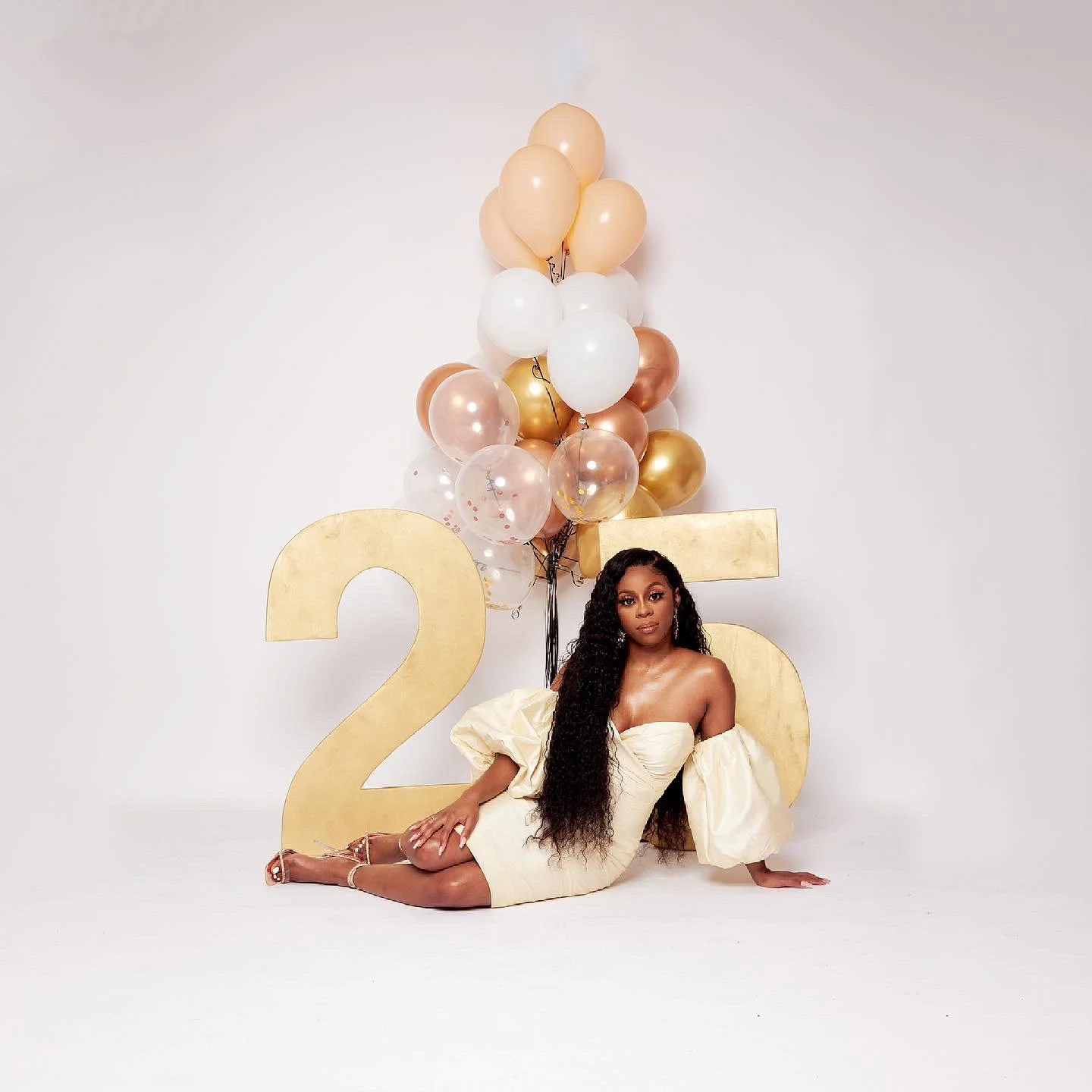 A woman in a cream-colored off-the-shoulder dress with puffy sleeves sitting on a white backdrop, surrounded by large gold and cream numbers '25' and decorated with a bunch of balloons in gold, white, and translucent with confetti inside, celebrating