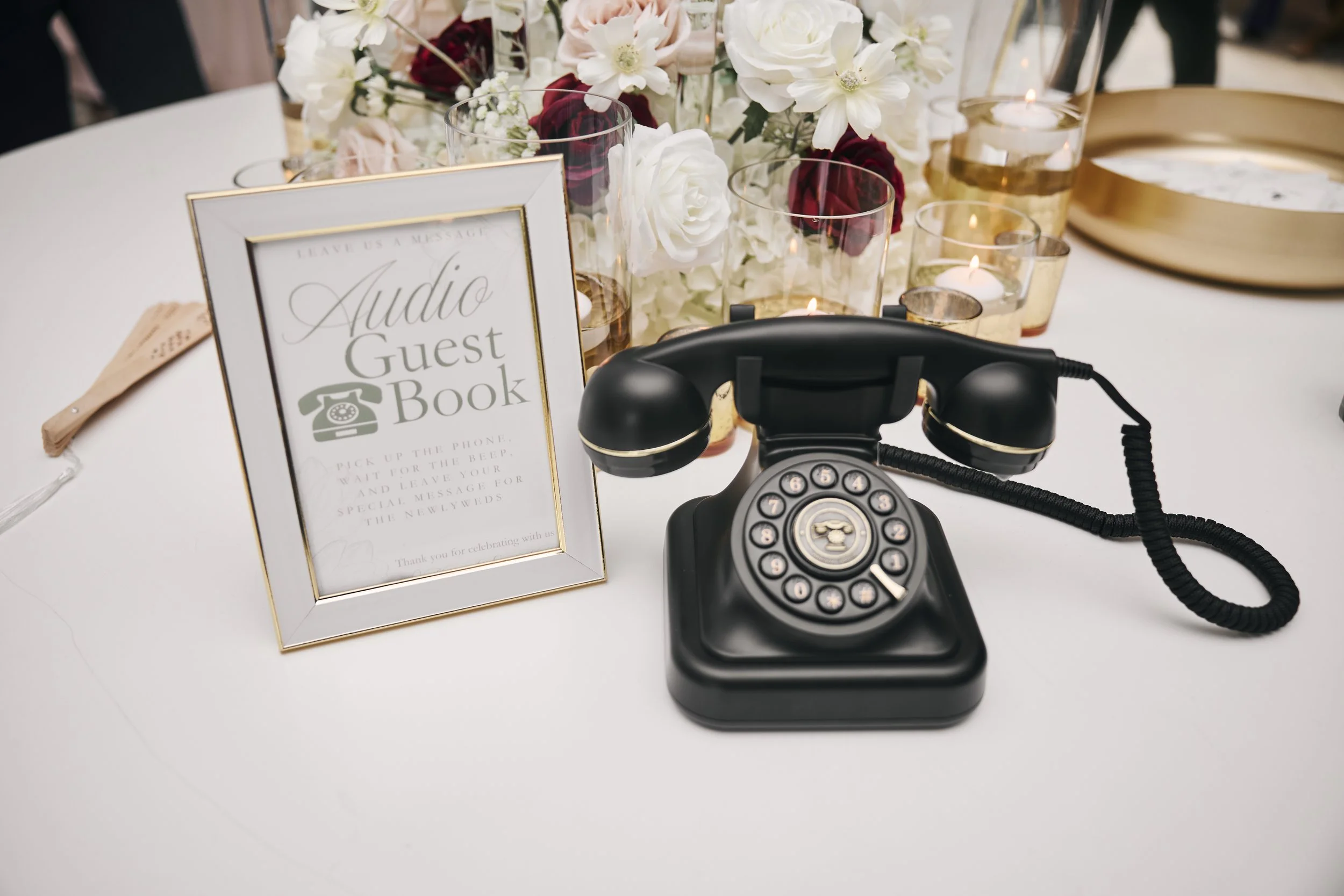 A vintage black rotary phone on a white table, surrounded by candles and a floral centerpiece, with a framed sign for an audio guest book at an event.