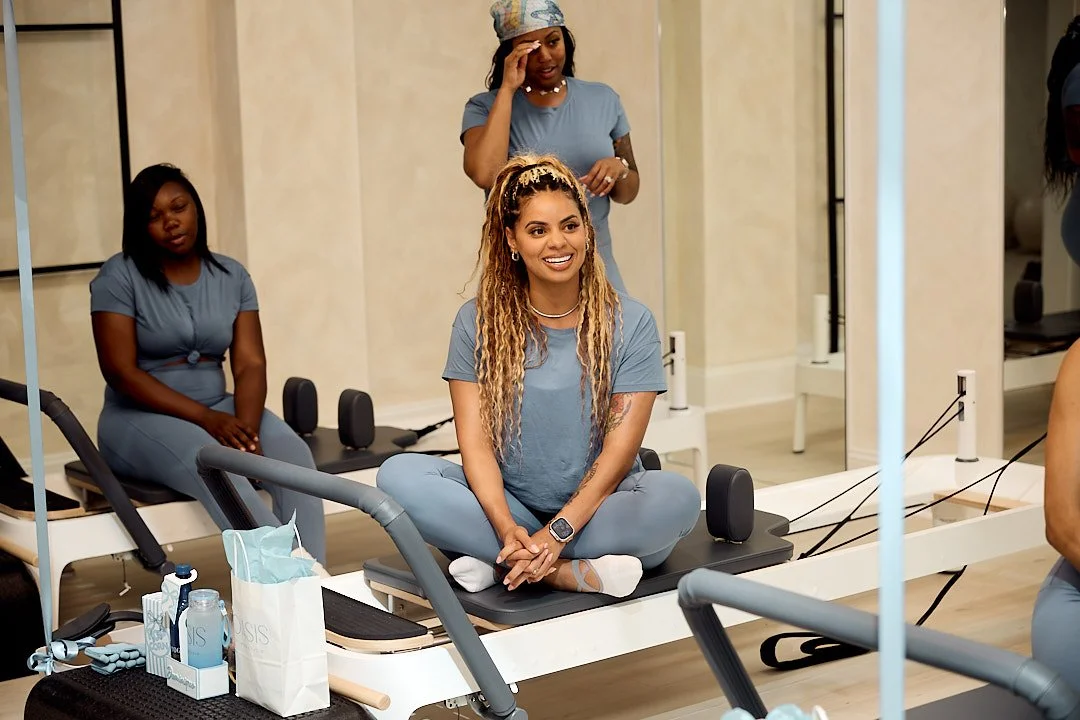 A group exercise class in a gym or fitness studio, featuring women sitting on reformer machines, with one woman in the foreground smiling and sitting cross-legged, while others are seated in the background. The women are wearing matching blue workout