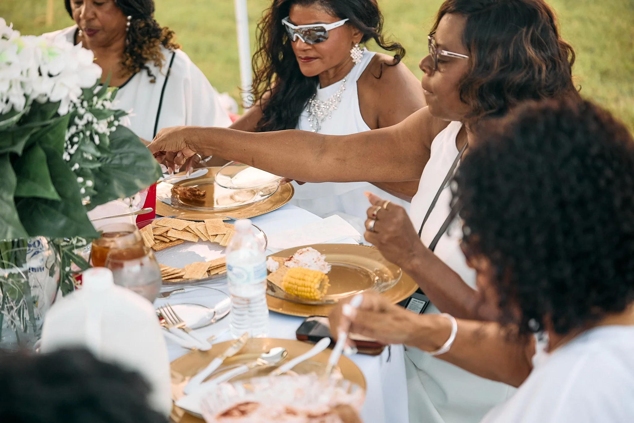 A group of women wearing white dresses and jewelry are sitting at a table outdoors, serving themselves food from a communal meal.
