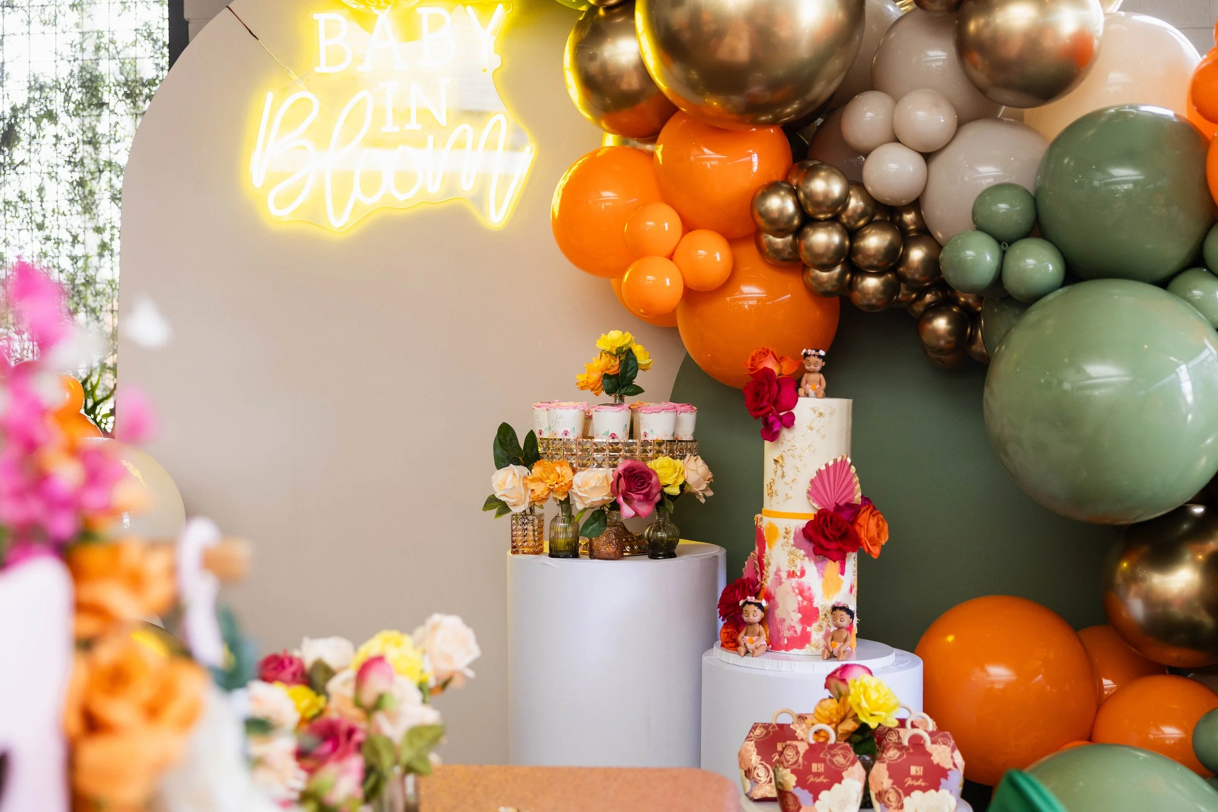 Decorative celebration setup with a balloon arch in orange, green, white, gold, and pink balloons. There are two tiered cakes with floral decorations, one with a pink fan and flowers, and others topped with small figurines of children. A neon sign in