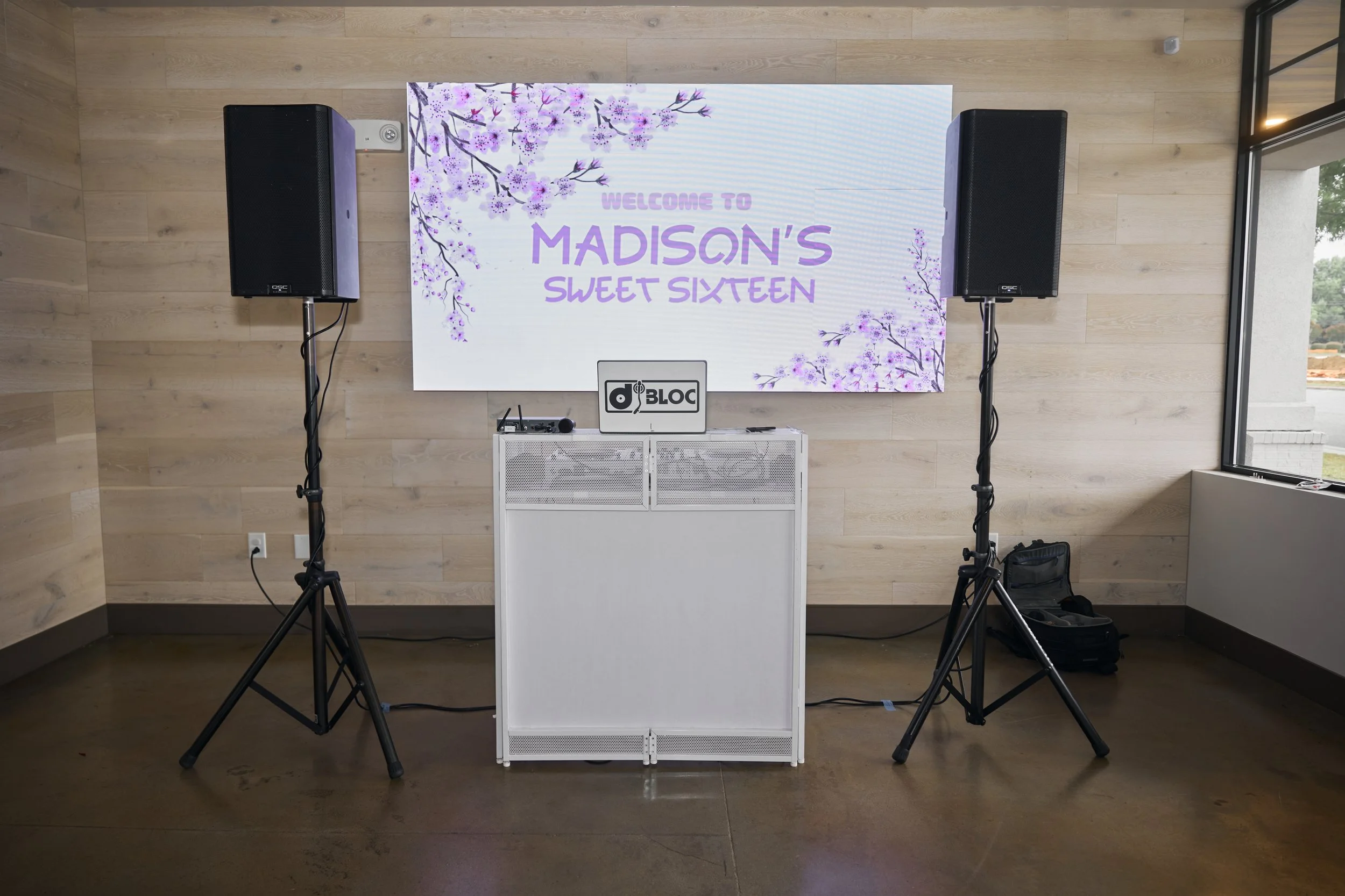 DJ setup with two black speakers on stands, a white DJ booth, and a digital screen displaying a welcome message for Madison's 16th birthday with cherry blossom graphics.