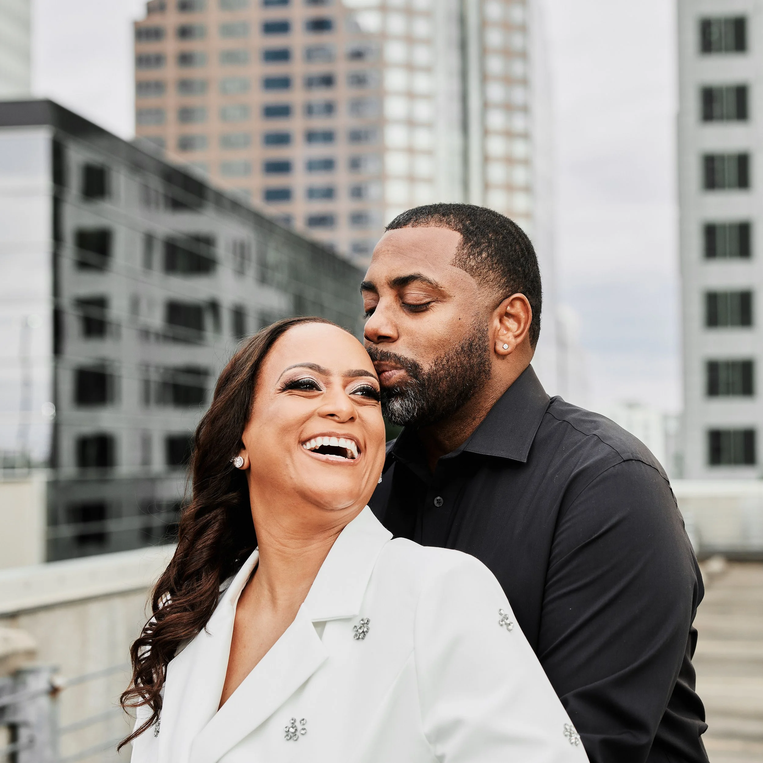 A happy couple standing close together outdoors in an urban setting, with tall buildings in the background. The woman is smiling and laughing, while the man is gently kissing her on the forehead.