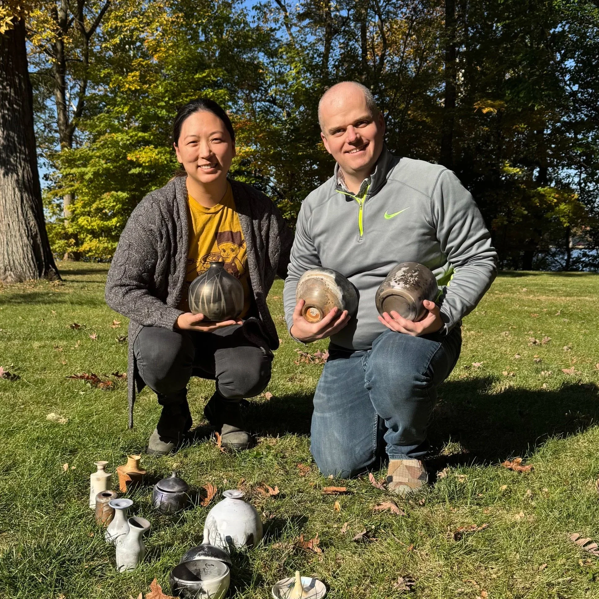 A man and woman kneeling outdoors on grass, holding ceramic pottery in front of a background of trees with autumn foliage.