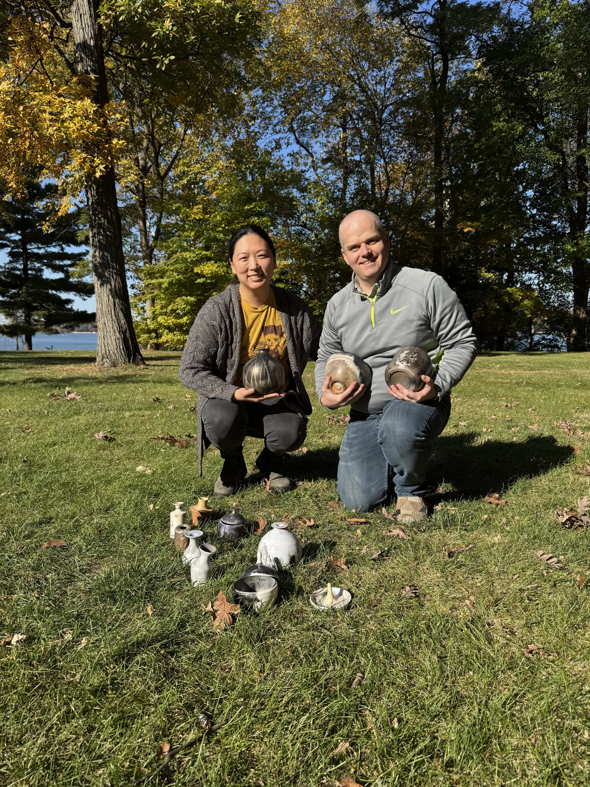 Two friends kneeling on grass in a park, holding ceramic vases, with more vases arranged on the ground in front of them, surrounded by trees with autumn leaves and a body of water in the background.