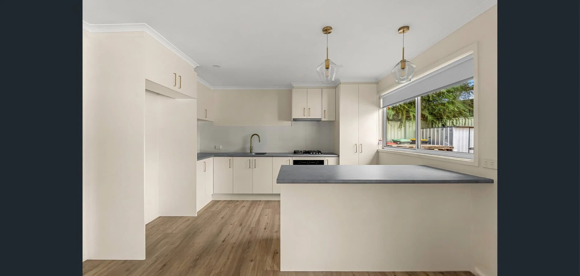 A modern, empty kitchen with white cabinets, gray countertops, a window with a view of a backyard with trees, and two hanging light fixtures.