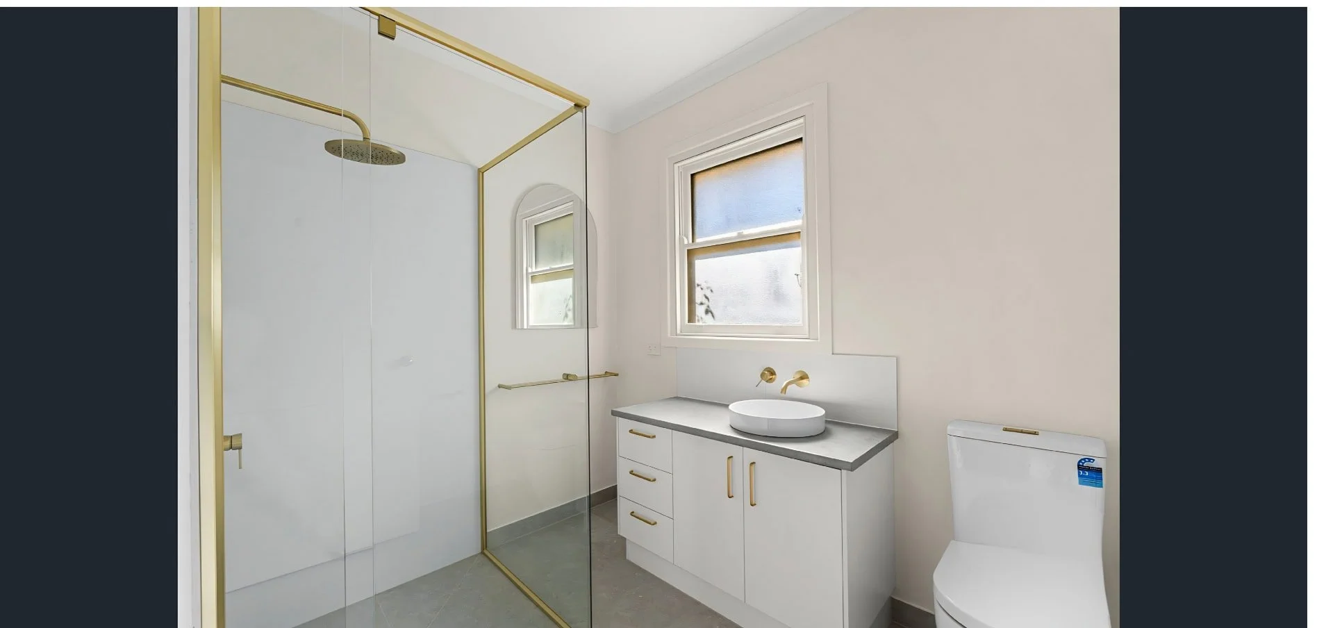Modern bathroom with white walls, a glass shower enclosure with gold trim, a white vanity with a round vessel sink, a large window, and a white toilet.