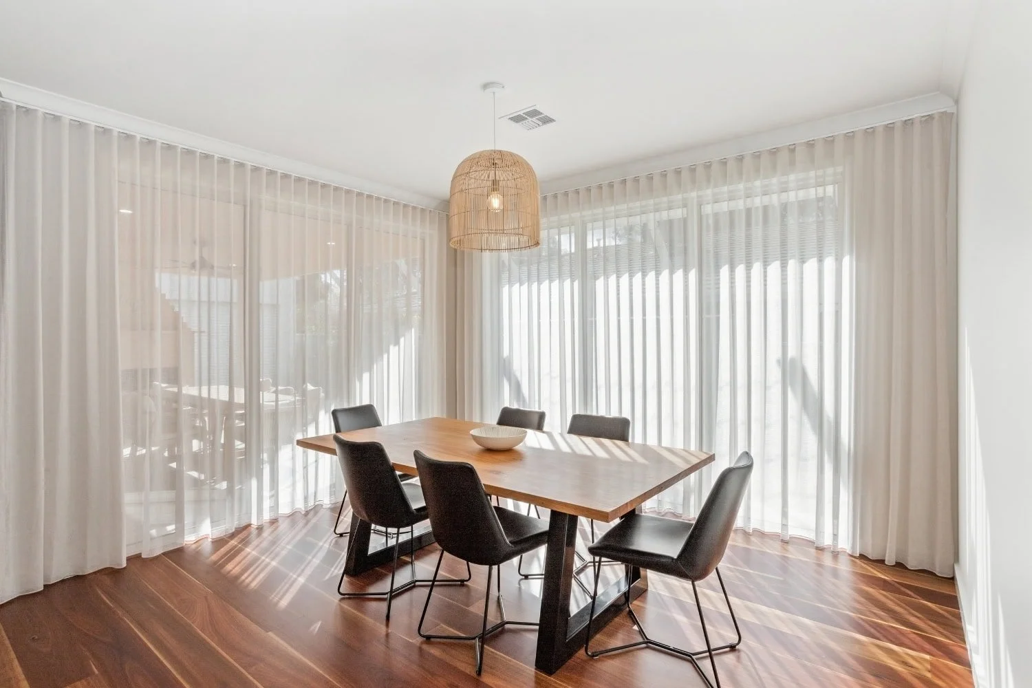 Light-filled dining room with a timber table, charcoal dining chairs and a woven rattan pendant, framed by sheer-curtained windows and warm timber flooring in a Marnie Rd Bendigo custom home.