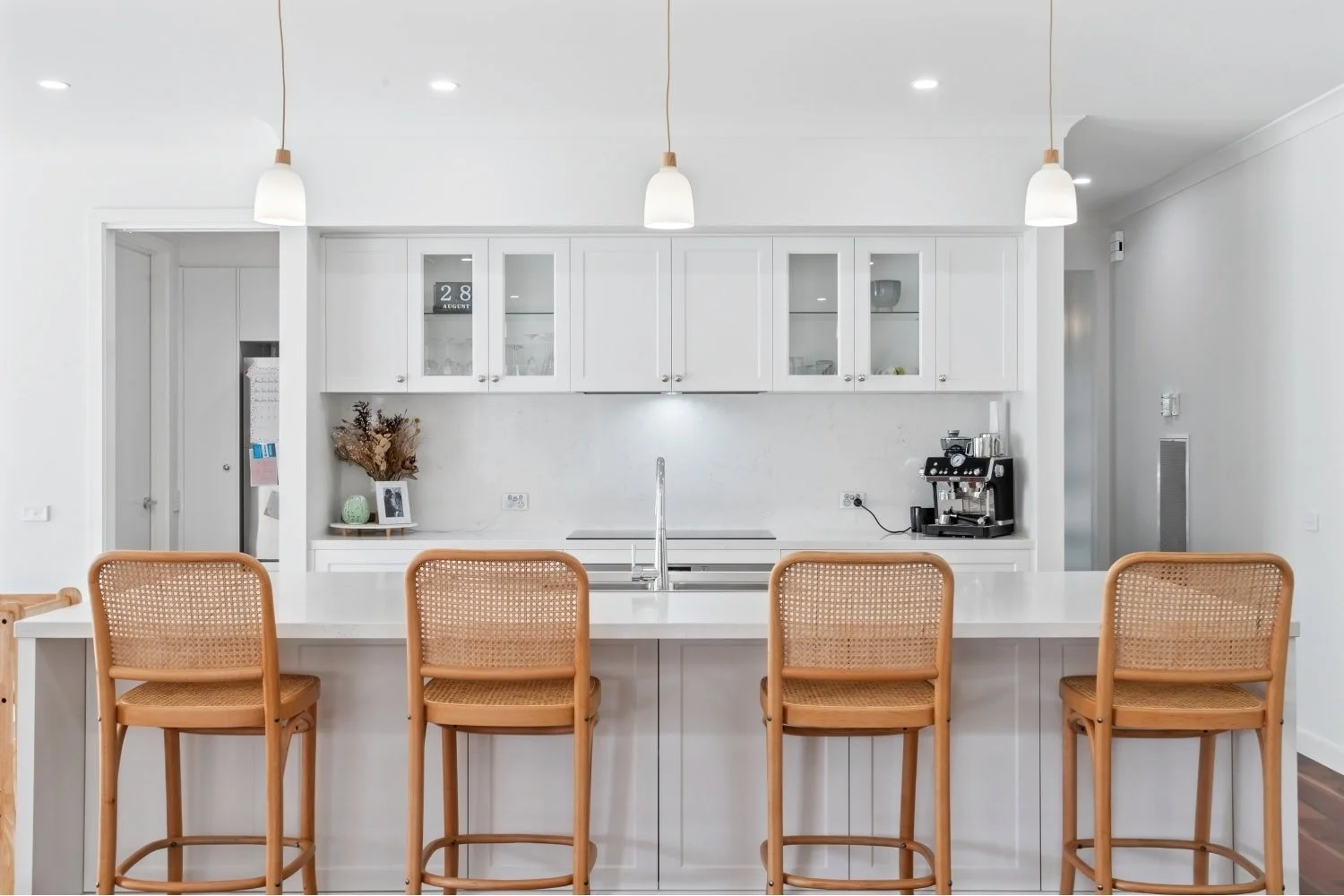 White shaker kitchen with stone island bench, cane bar stools, glass-front cabinetry, and timber pendant lighting at Marnie Road, Bendigo, by AMW Builders.