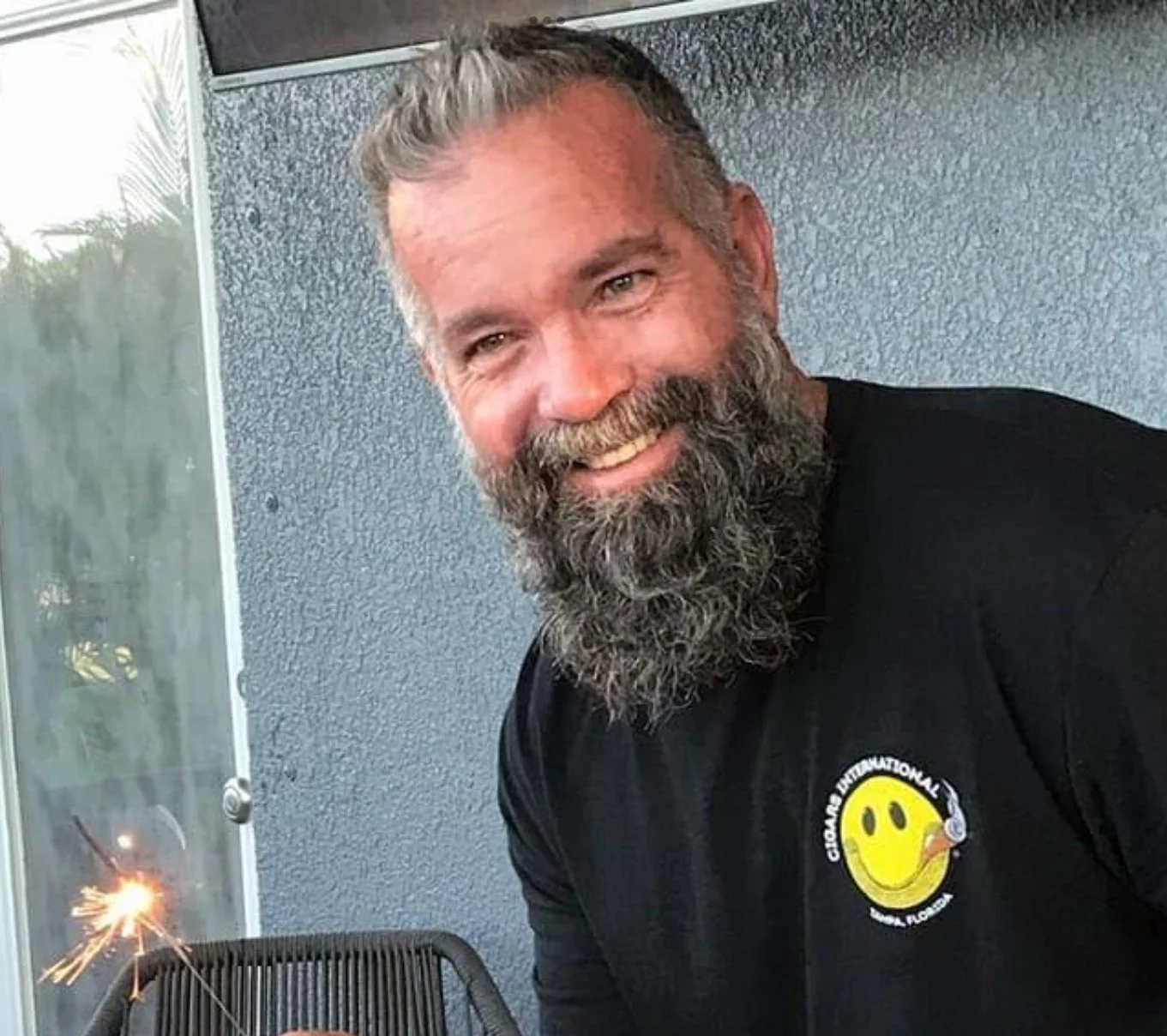 A man with gray hair and a full beard smiling at the camera, wearing a black shirt with a yellow smiley face logo on it, sitting outside near a gray wall, with a lit sparkler in front of him.