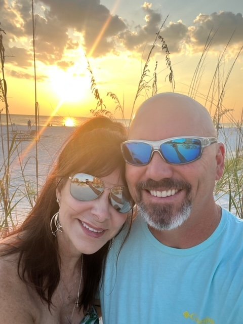 A smiling couple taking a selfie on the beach at sunset, with tall grass in the foreground and the ocean in the background, both wearing sunglasses.