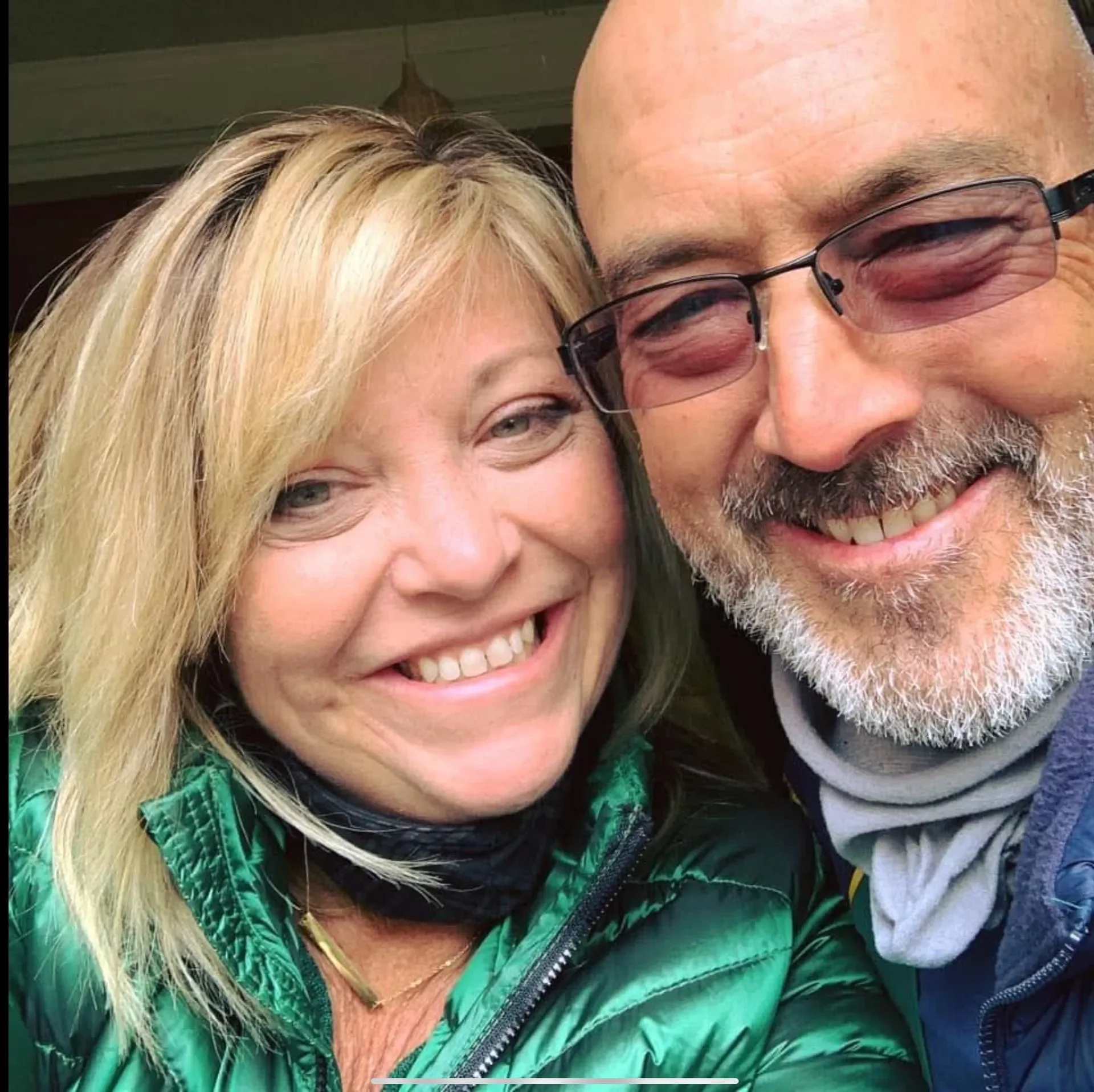 A smiling woman with blonde hair and a woman with glasses, a beard, and a gray scarf taking a close-up selfie together.