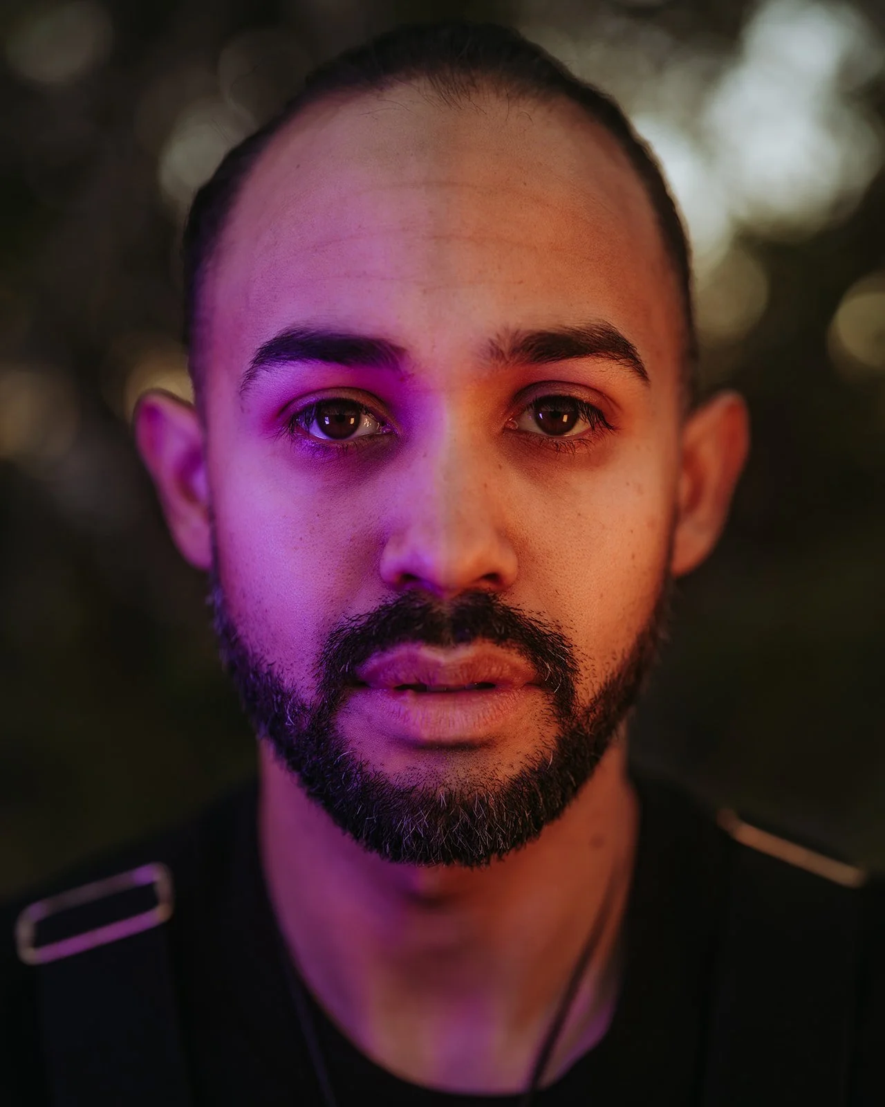 Portrait of a man with a beard, lit by purple and orange lights, against a blurred background.