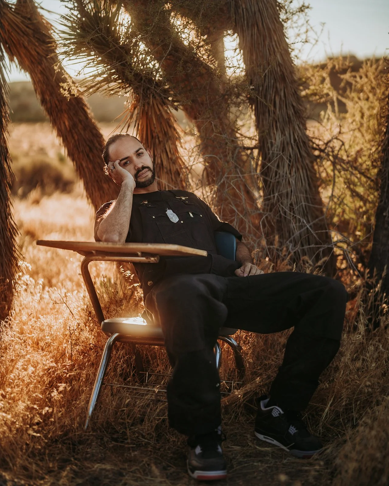 Person sitting in a school desk chair outdoors near trees, in a dry grassy field, with sunlight filtering through branches.