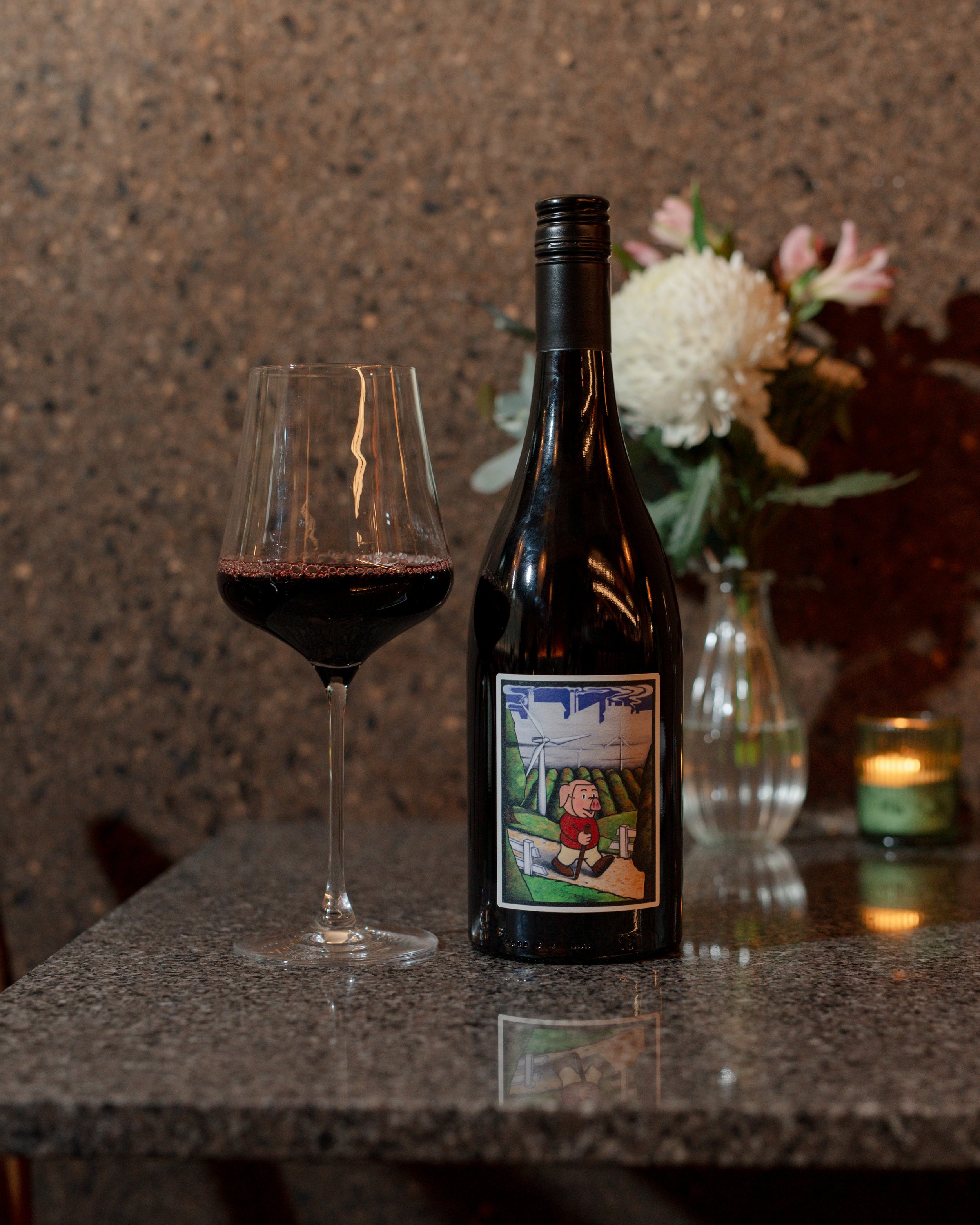A wine glass partially filled with red wine, a wine bottle with a colorful label depicting a cartoon man with a walking stick, and a background with a floral arrangement and candles on a stone surface.