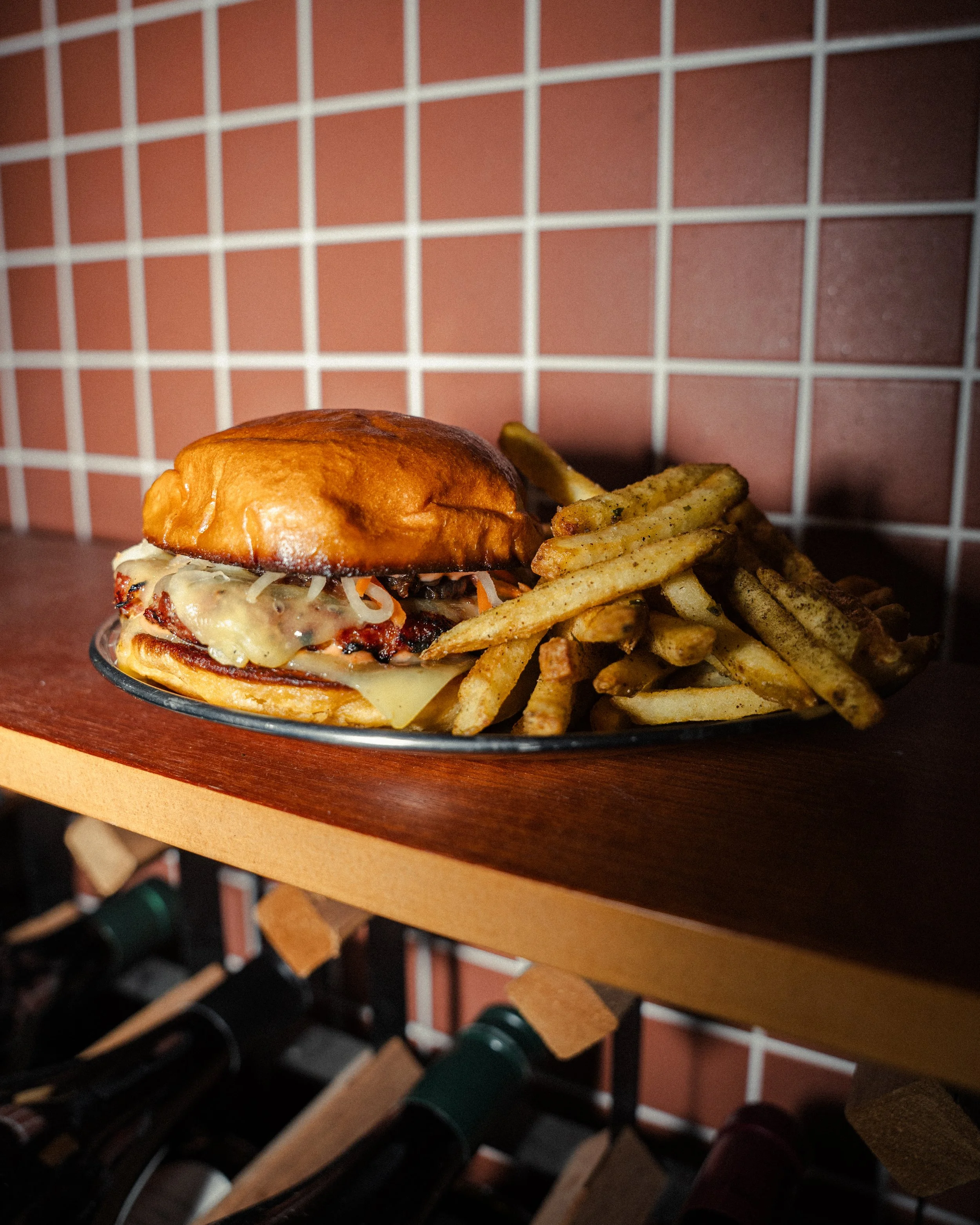 A Chori Burger with melted cheese vserved with French fries on a metal tray, with a pink tiled wall in the background.