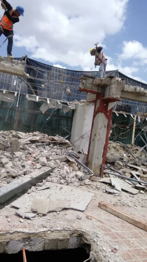 Dos trabajadores con cascos trabajando en una estructura de construcción o demolición en un sitio lleno de escombros y materiales rotos.