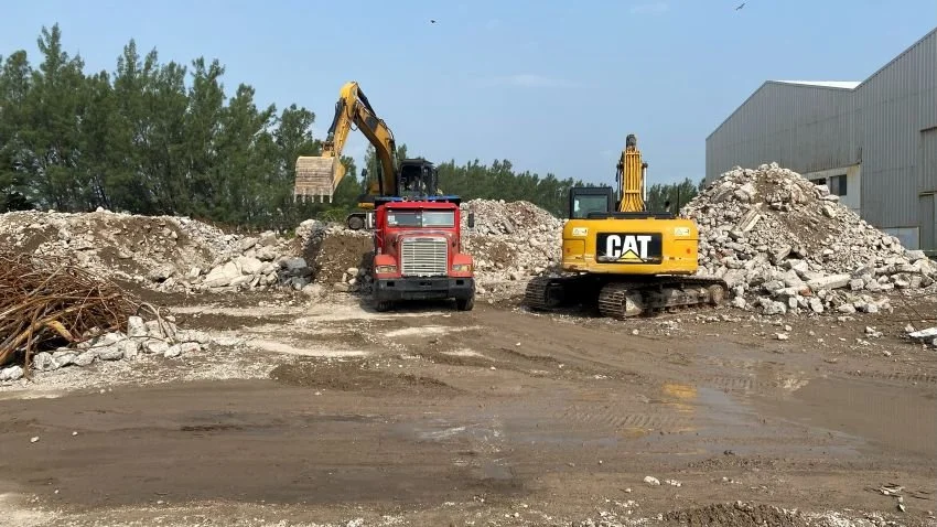 Maquinaria pesada en un sitio de construcción, con excavadora amarilla y camión rojo, trabajando en tierra y escombros, cerca de un almacén o estructura industrial.
