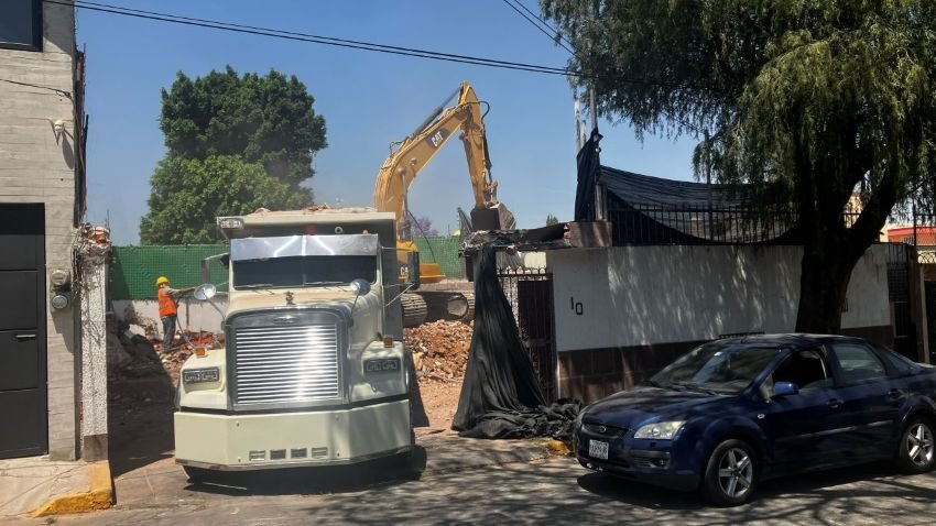 Maquinaria de construcción y una camioneta en una obra en una calle con árboles y un coche negro estacionado.