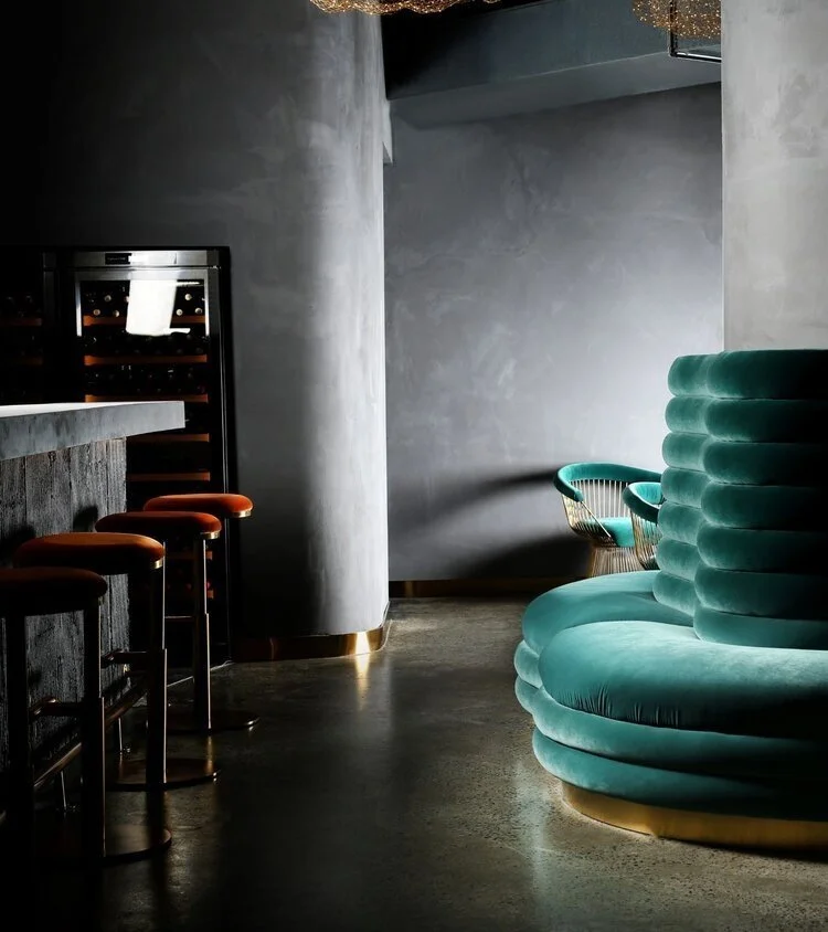 Interior of a modern bar with a concrete floor, a curved green velvet seating area, and a small bar counter with four barstools. There is a wine cooler against the wall, and a small round table with green chairs in the background.