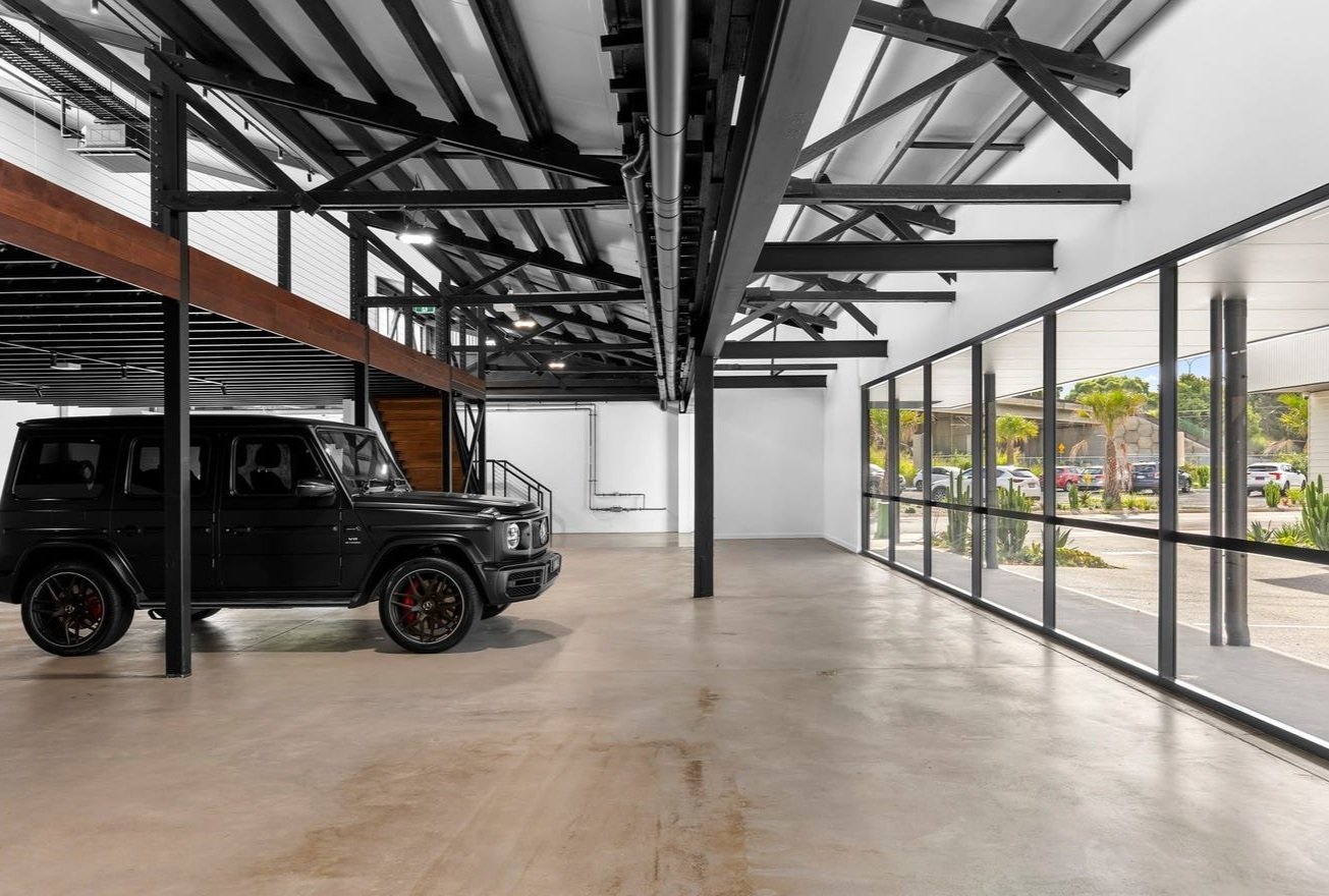 Empty indoor showroom or garage with a black vehicle parked and large glass windows showing outside parking lot and plants.
