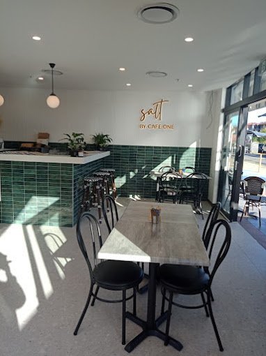 Interior of a modern cafe with a wooden table and black chairs near large glass windows and green tiled counter with plants, sunny with shadows cast on the floor.