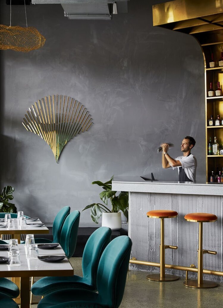 Modern restaurant interior with teal chairs at a dining table, a bar area with gold stools, a bartender shaking a cocktail, and decorative gold wall art and shelving.