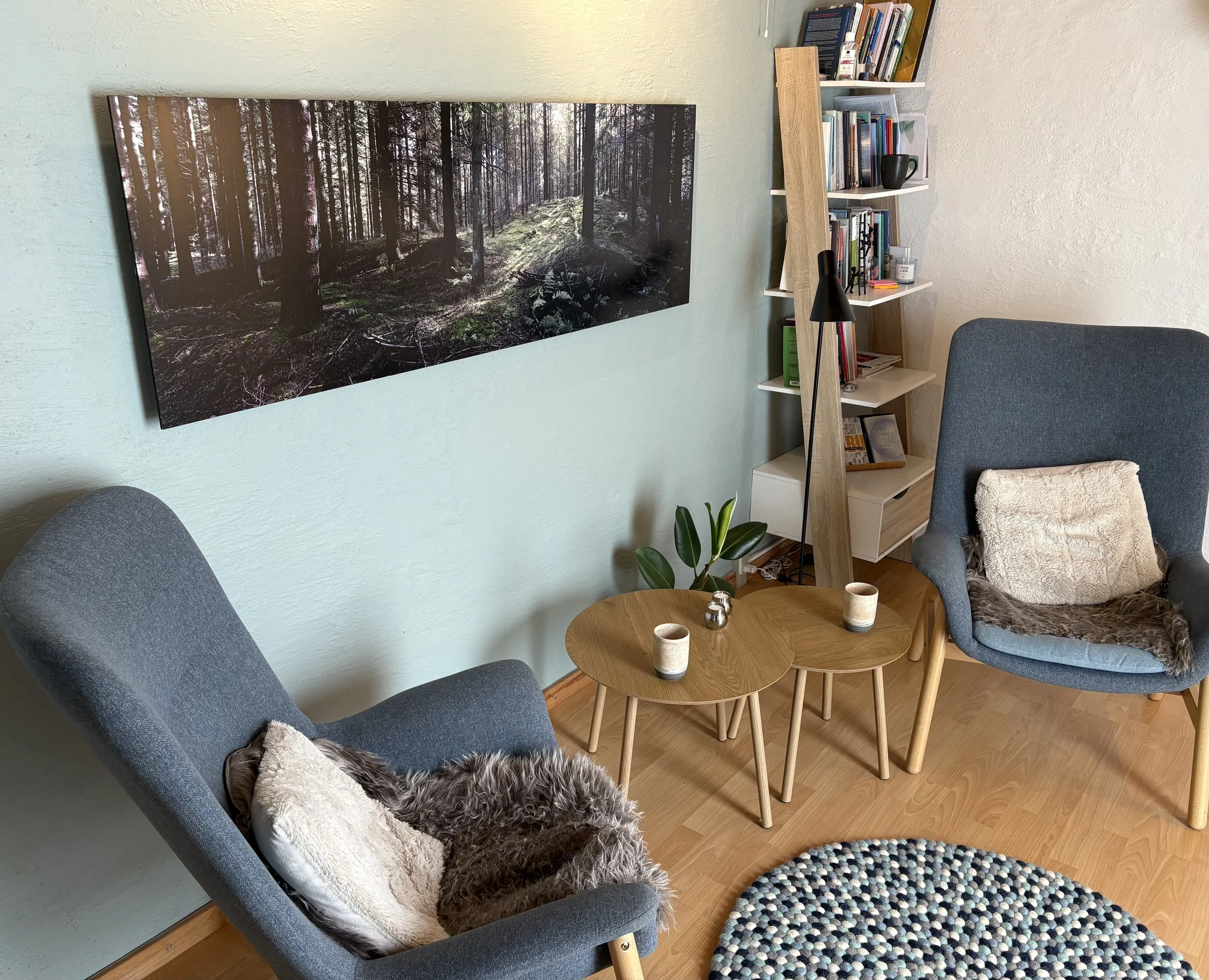 Et hyggeligt stuebillede med to blå armlæn sofaer, et rundt træbord med tre kopper, en plante, en moderne gulvlampe, en reol fyldt med bøger, en stol med en fluffy pude, og et stort vægbillede af en skov.