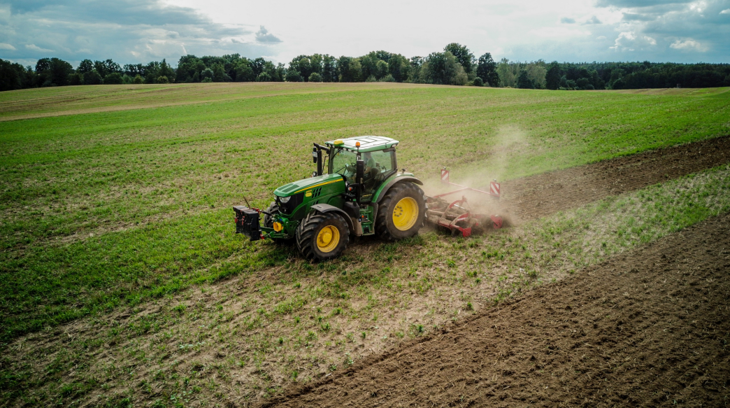 A green tractor working on a large farm field under a cloudy sky.