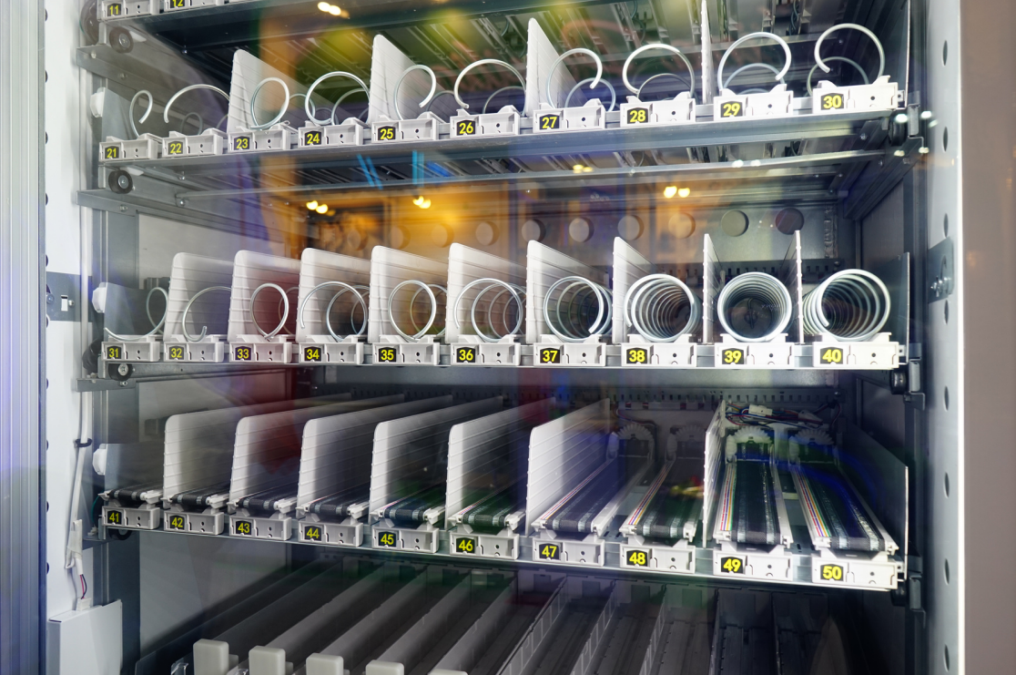 A vending machine with various coiled and straight charging cables and cords organized on shelves, numbered from 21 to 50.
