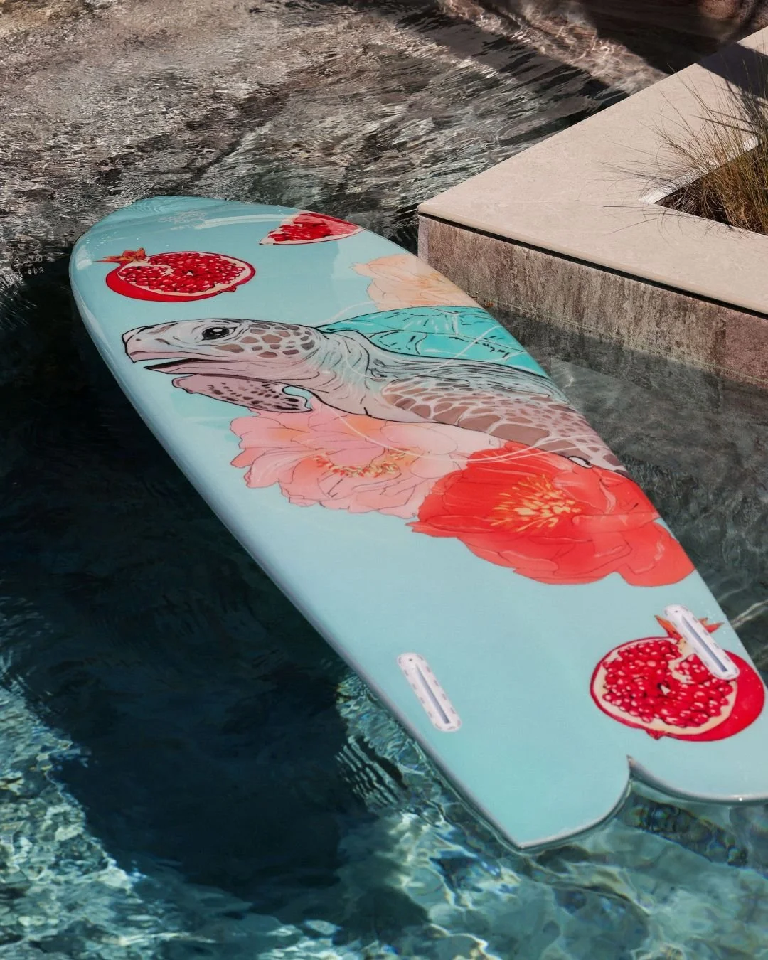 Planche de paddle avec des illustrations de fleurs, d’un lézard, et de grenades, flottant dans une piscine.