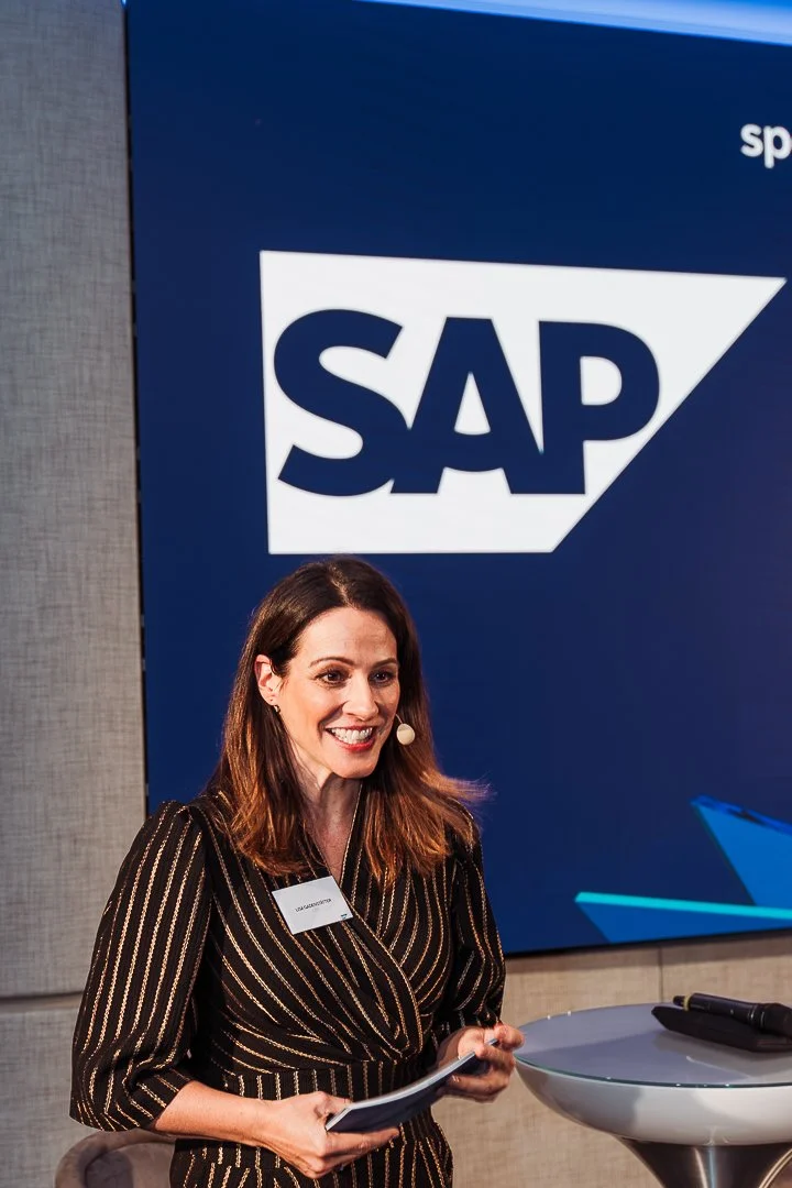 A woman standing in front of a large SAP logo on a blue background, smiling, holding documents, and wearing a striped black and gold dress.