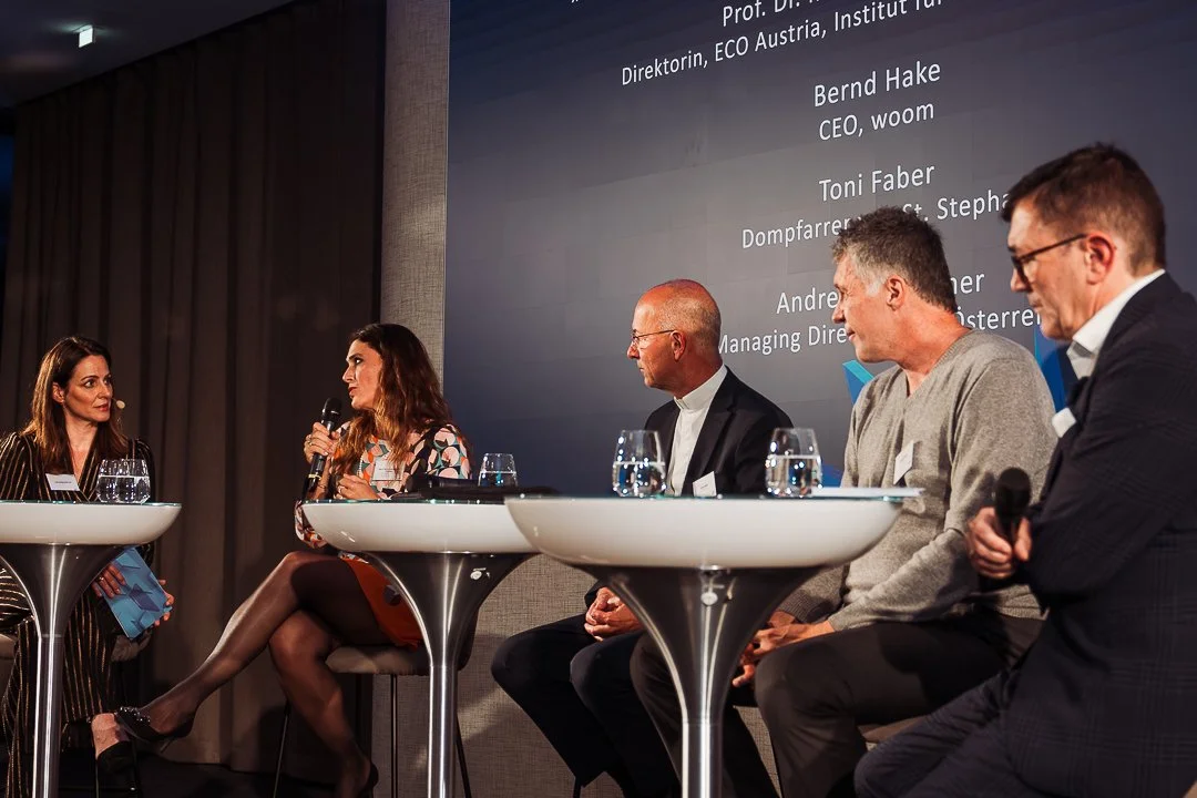 A panel discussion with five people on stage in front of a large screen, four men and one woman, all seated behind white round tables with glasses of water. The woman is speaking into a microphone, and the man on the far right is also holding a microphone. The background shows names and titles of the speakers on the screen.