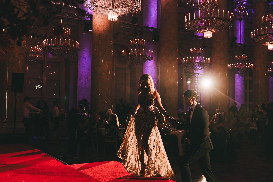 Vienna Awards!A woman in a glamorous gown and a man in a suit dance on a red carpet at an elegant, chandeliers-lit event with purple lighting.