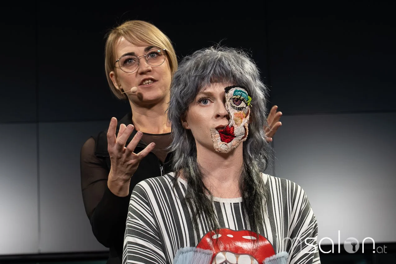 Imsalon.Two women on stage, one with short blonde hair and glasses speaking with a microphone, the other with long gray hair wearing a patchwork face mask and striped shirt with a red tongue graphic, against a black background.