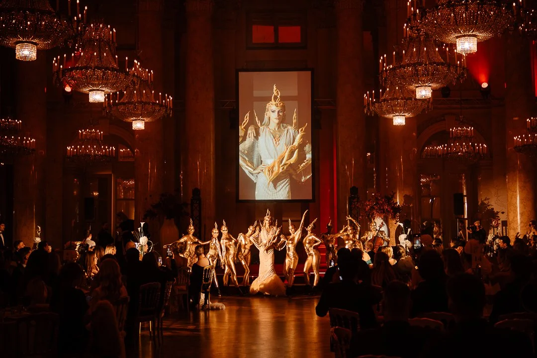 Vienna Awards.A performance with dancers dressed in gold costumes on a stage in an elegant, chandelier-lit ballroom. An audience watches as a large screen behind shows a close-up of a woman in a regal costume.